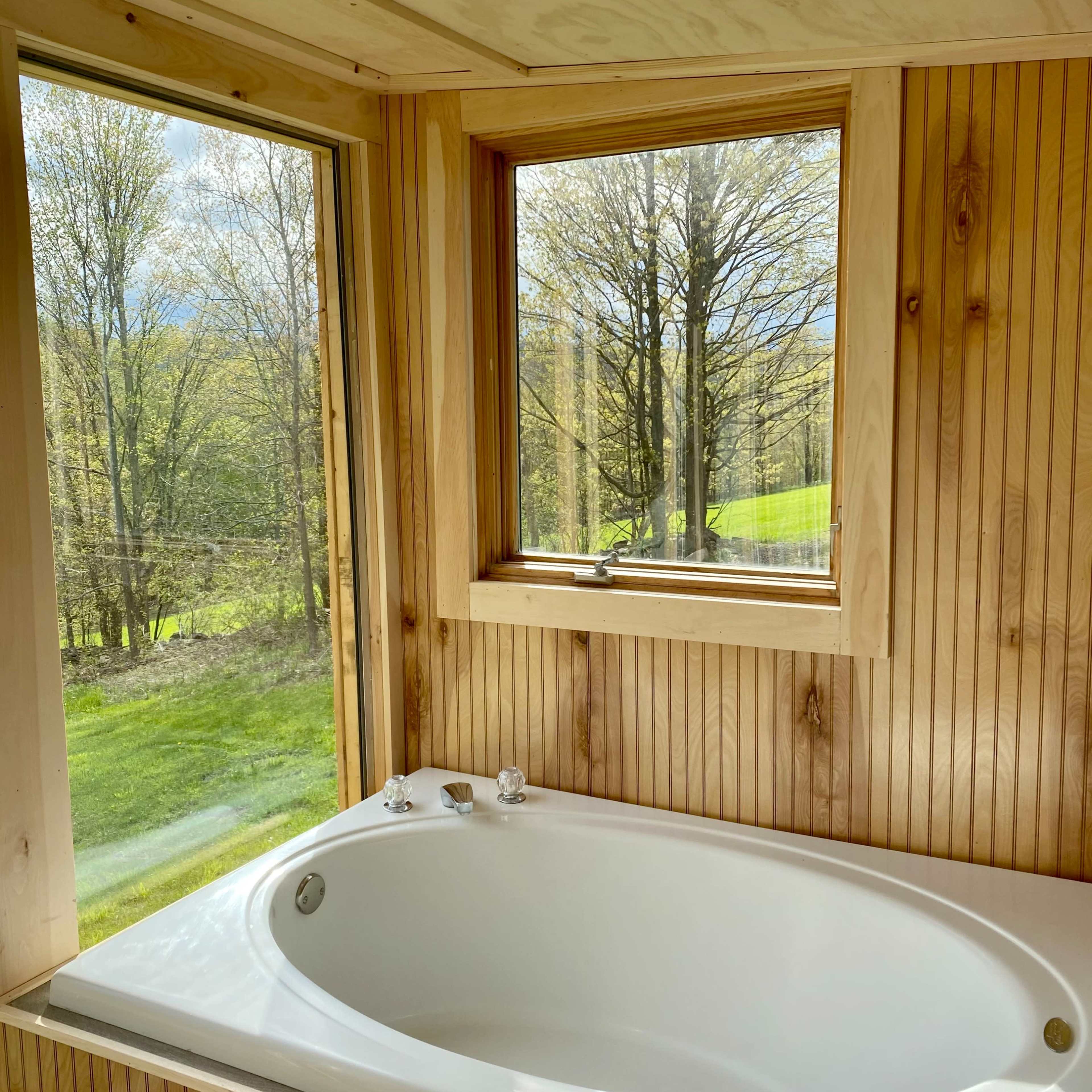 A white bathtub is situated in a wooden bathroom with large windows showcasing a view of trees and grassy landscape outside.