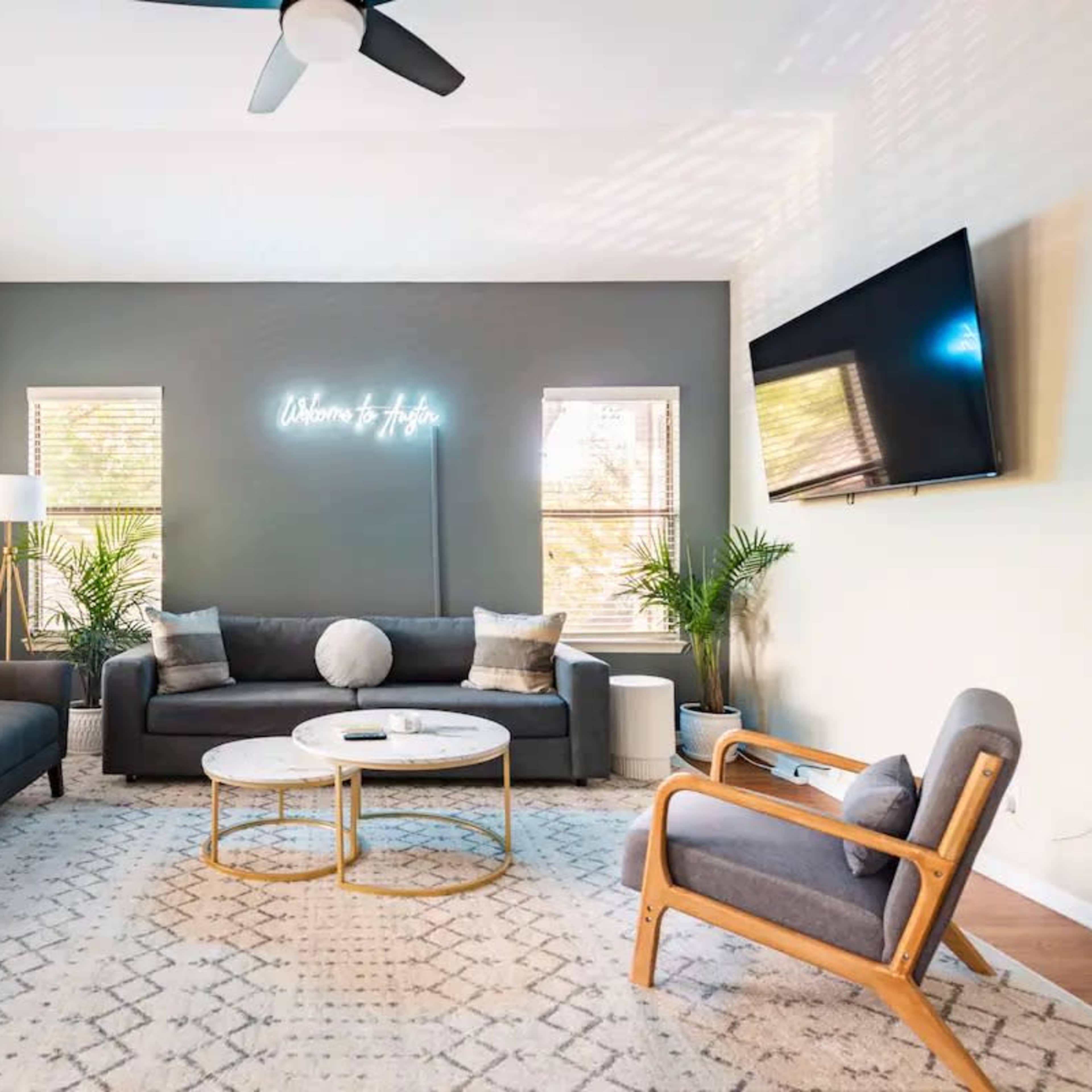 The living room features two dark sofas, a round coffee table, and a television mounted on the wall, with large windows allowing natural light to enter.