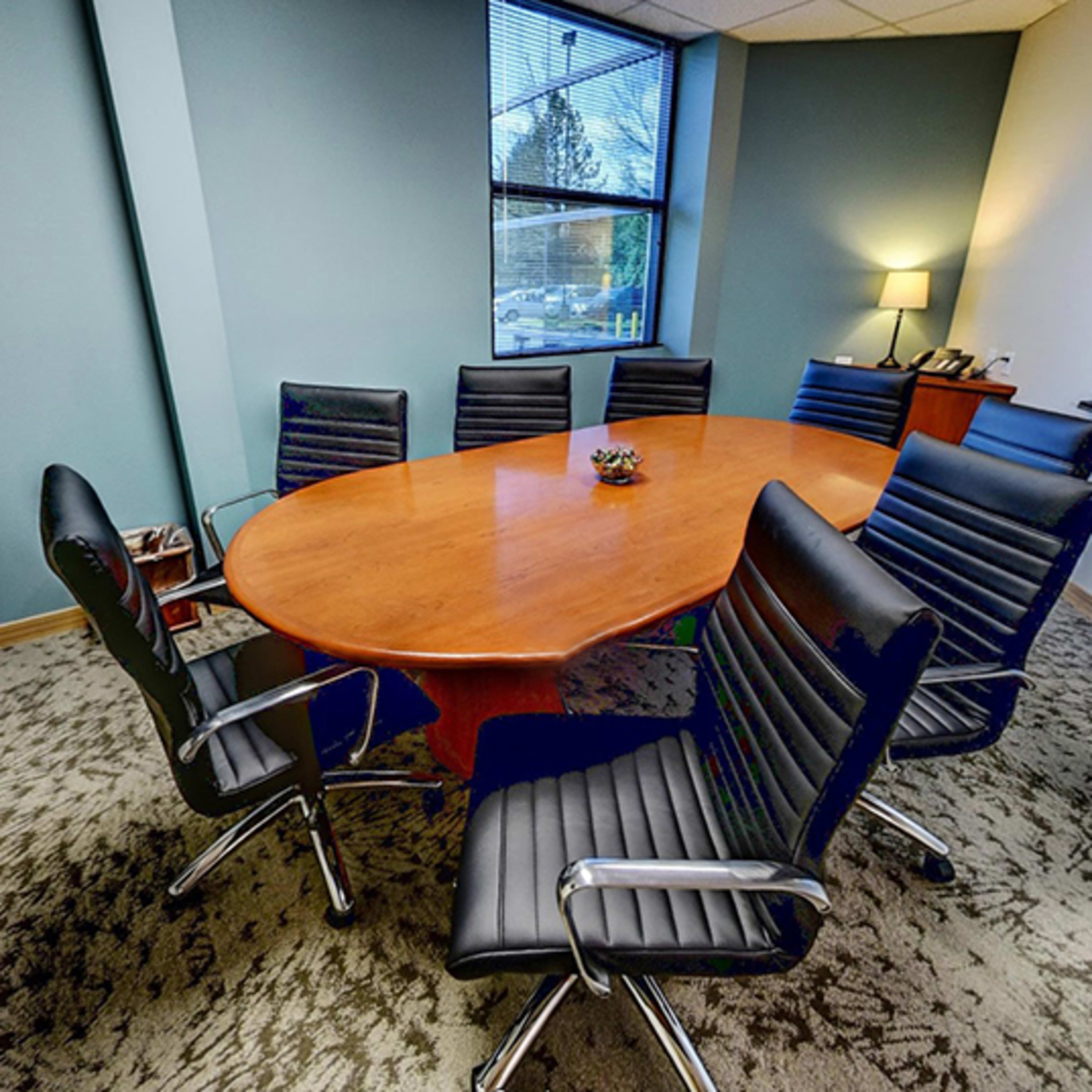 A conference room features an oval wooden table surrounded by black leather chairs, with a vase and a small plant on the table.
