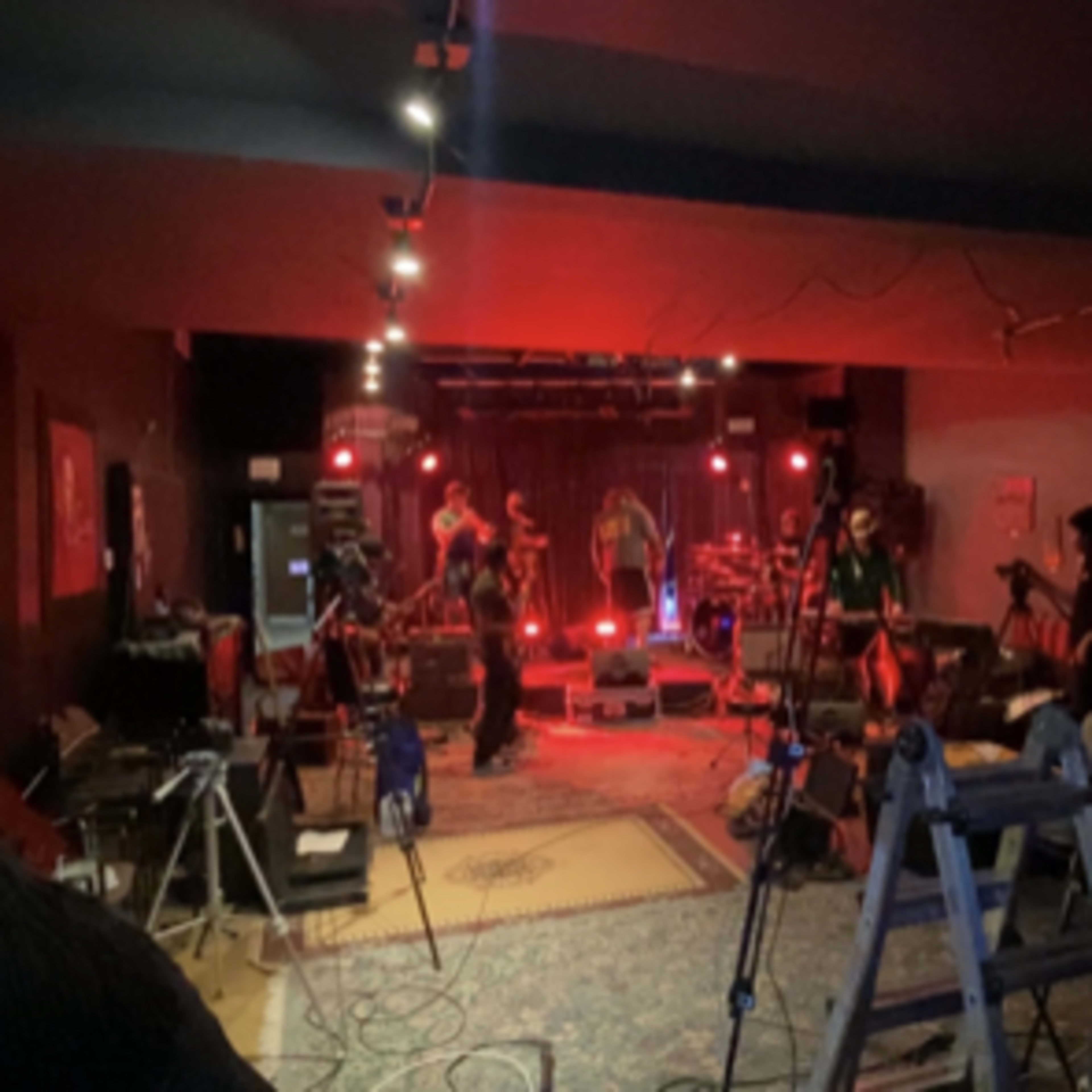 A dimly lit music venue set up for a performance, featuring red stage lights, musical instruments, and equipment arranged on the floor.