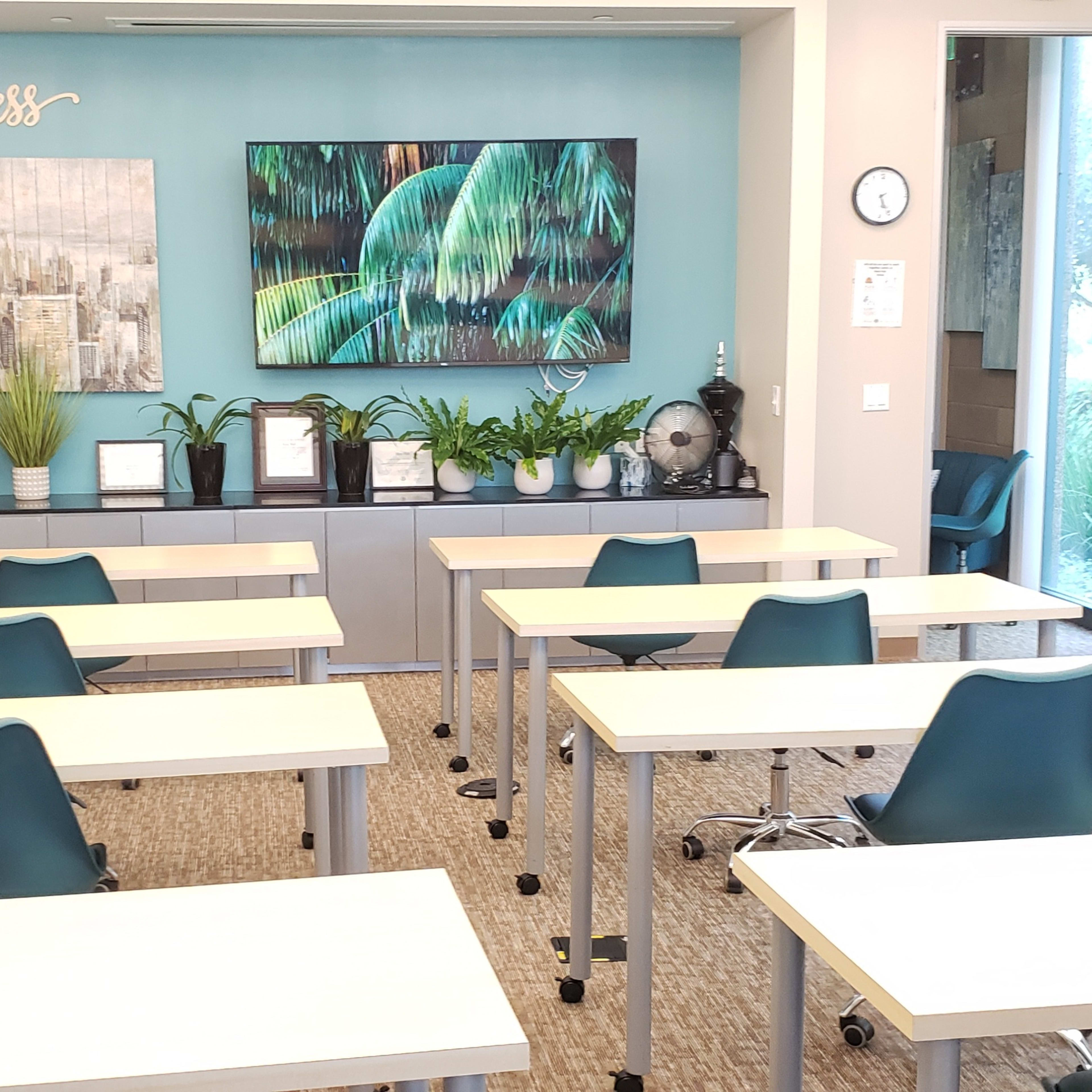 A modern classroom features several white tables and teal chairs arranged facing a wall adorned with two large artworks and potted plants, with a whiteboard on the left side.
