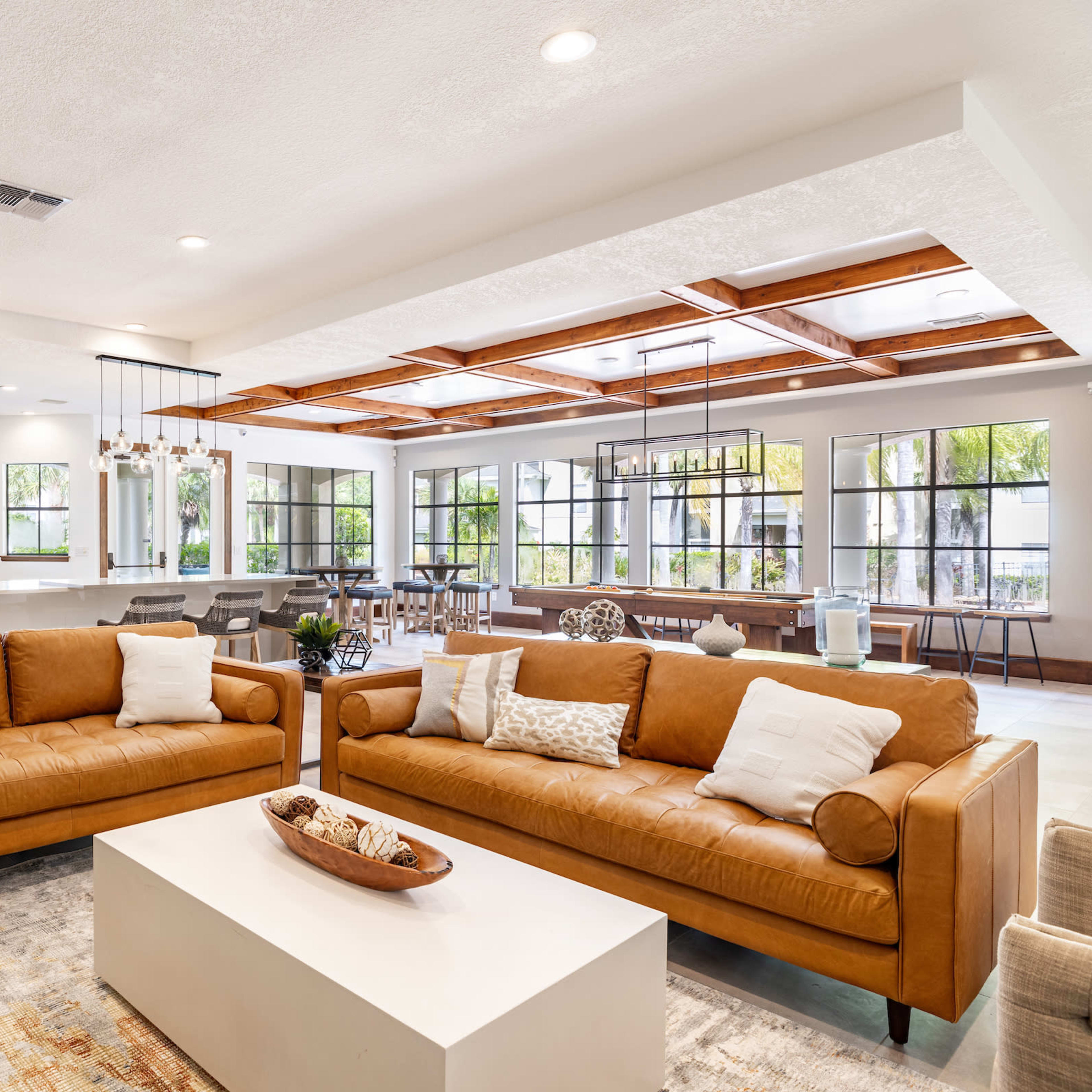 A modern living room furnished with two brown leather sofas facing a white coffee table, surrounded by bright windows and an open dining area in the background.