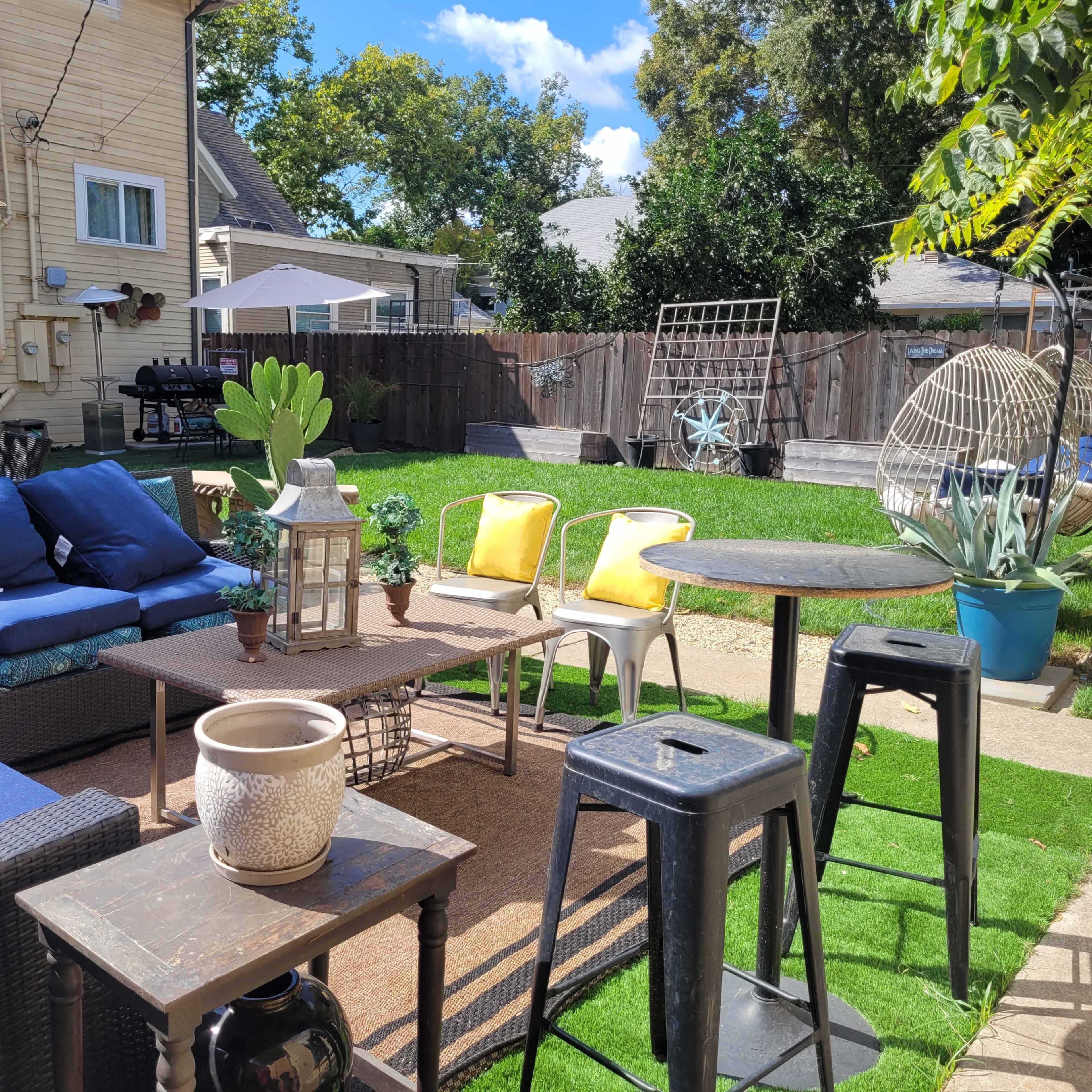 A backyard patio with outdoor furniture, including a seating area with blue cushions, a table, and a grassy area surrounded by wooden fences and various potted plants.