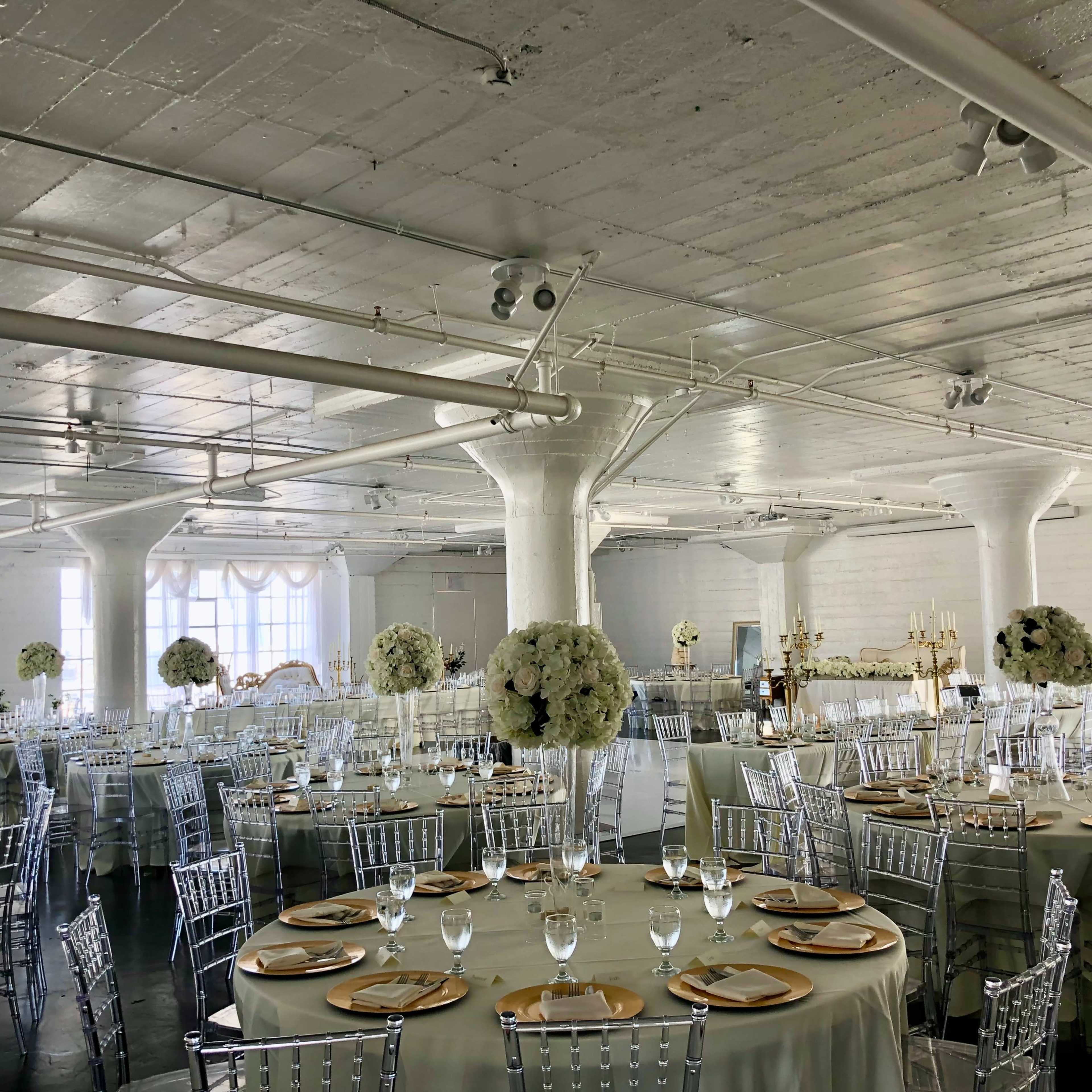 A spacious event hall is set with round tables covered in light-colored tablecloths, adorned with floral centerpieces and surrounded by clear acrylic chairs.