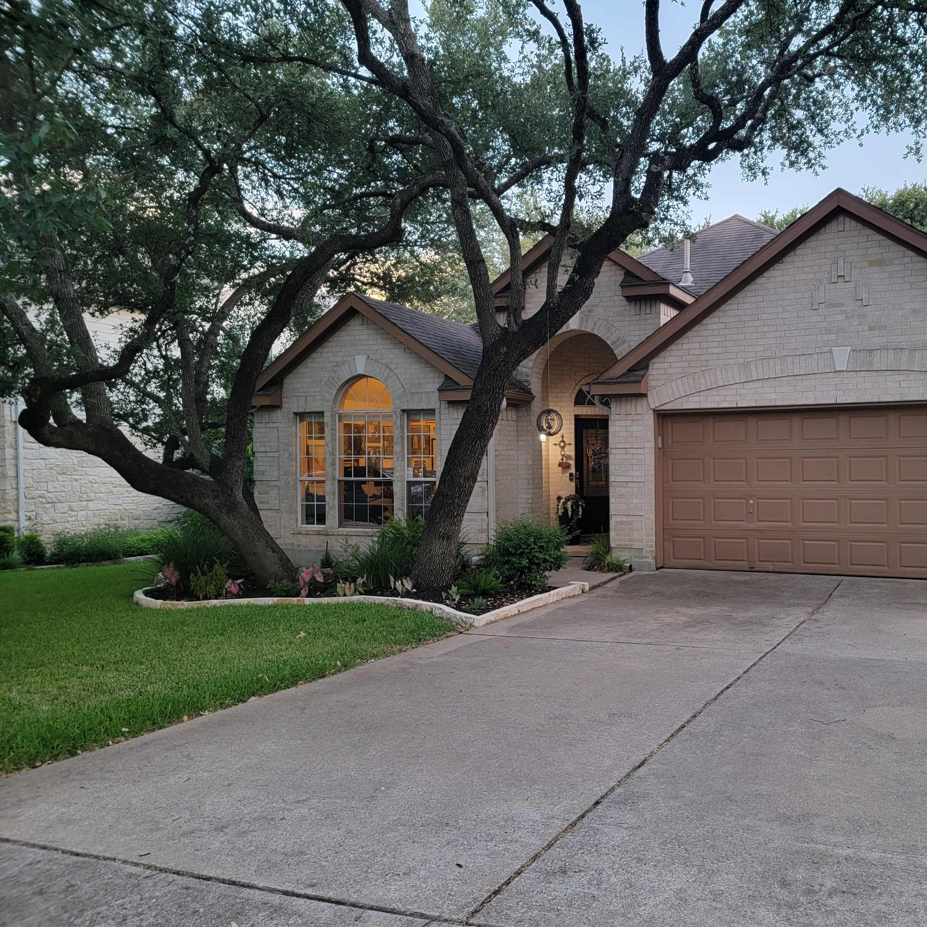 A single-story brick house with a garage sits on a neatly landscaped lot, featuring large trees and a well-maintained lawn.