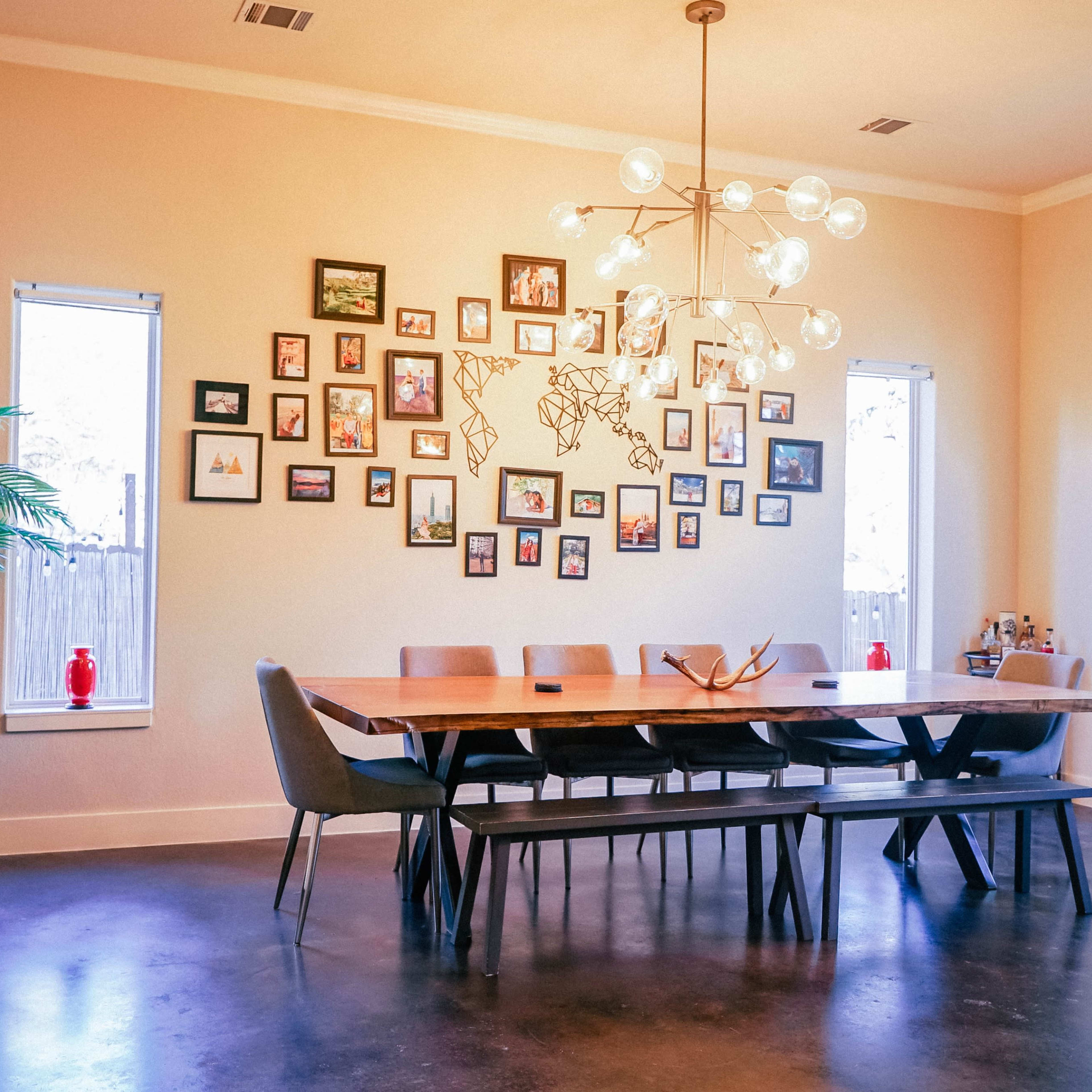 A spacious dining room features a long wooden table surrounded by gray chairs, with a gallery of framed photos on the wall and large windows letting in natural light.