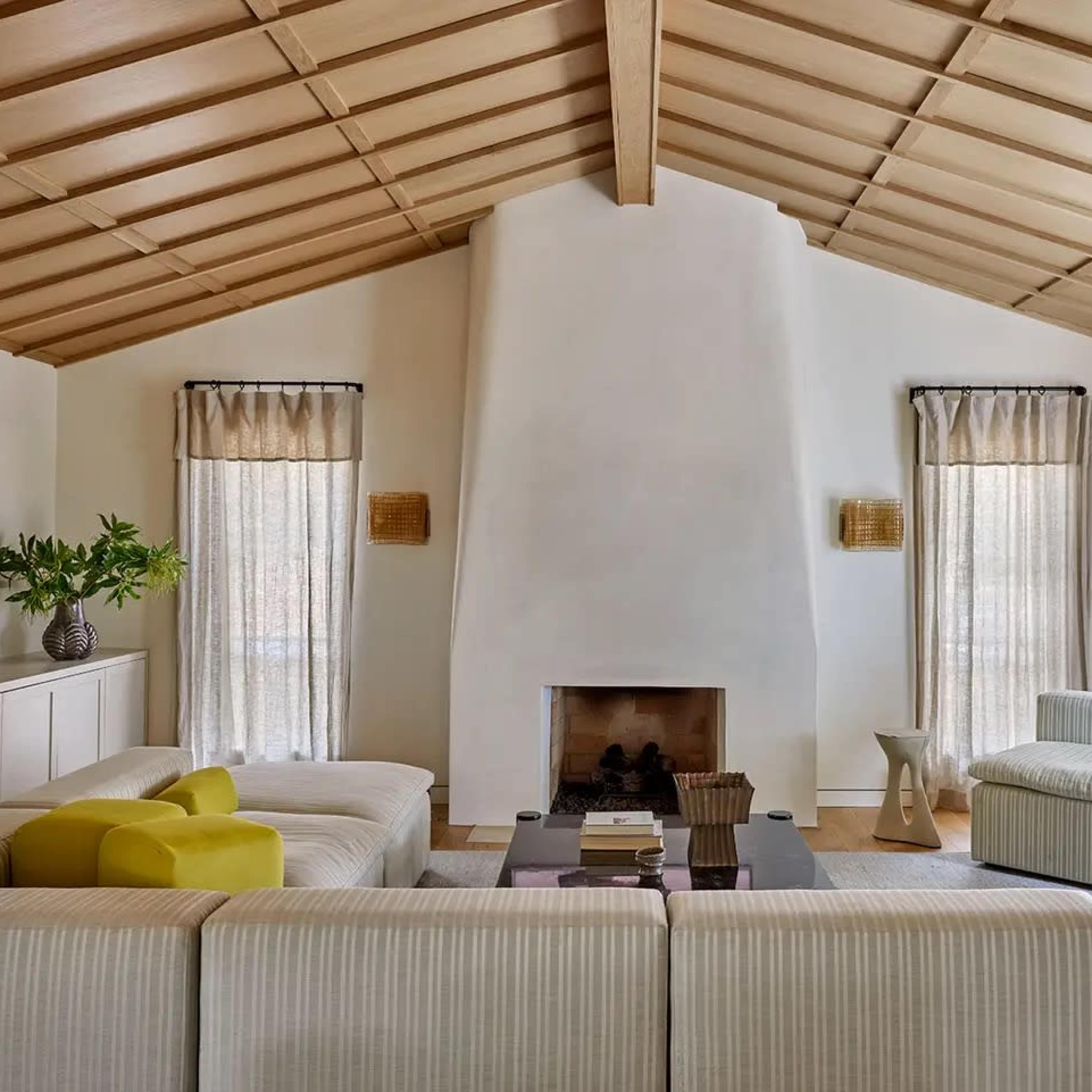 A spacious living room features a white fireplace and large windows draped with sheer curtains, surrounded by a sectional sofa and modern furniture.