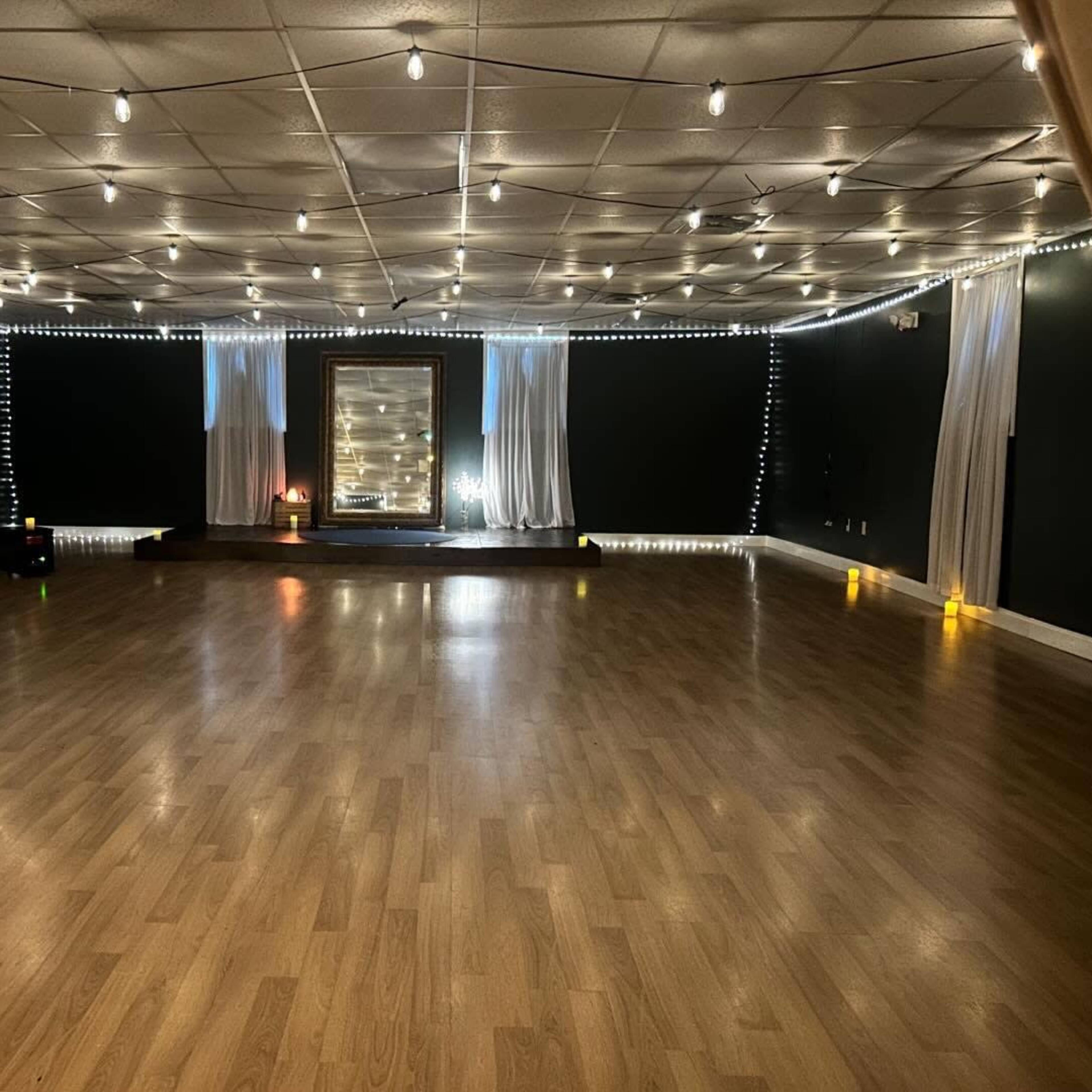 A spacious room with wooden flooring, decorated with string lights and a decorative backdrop at one end.