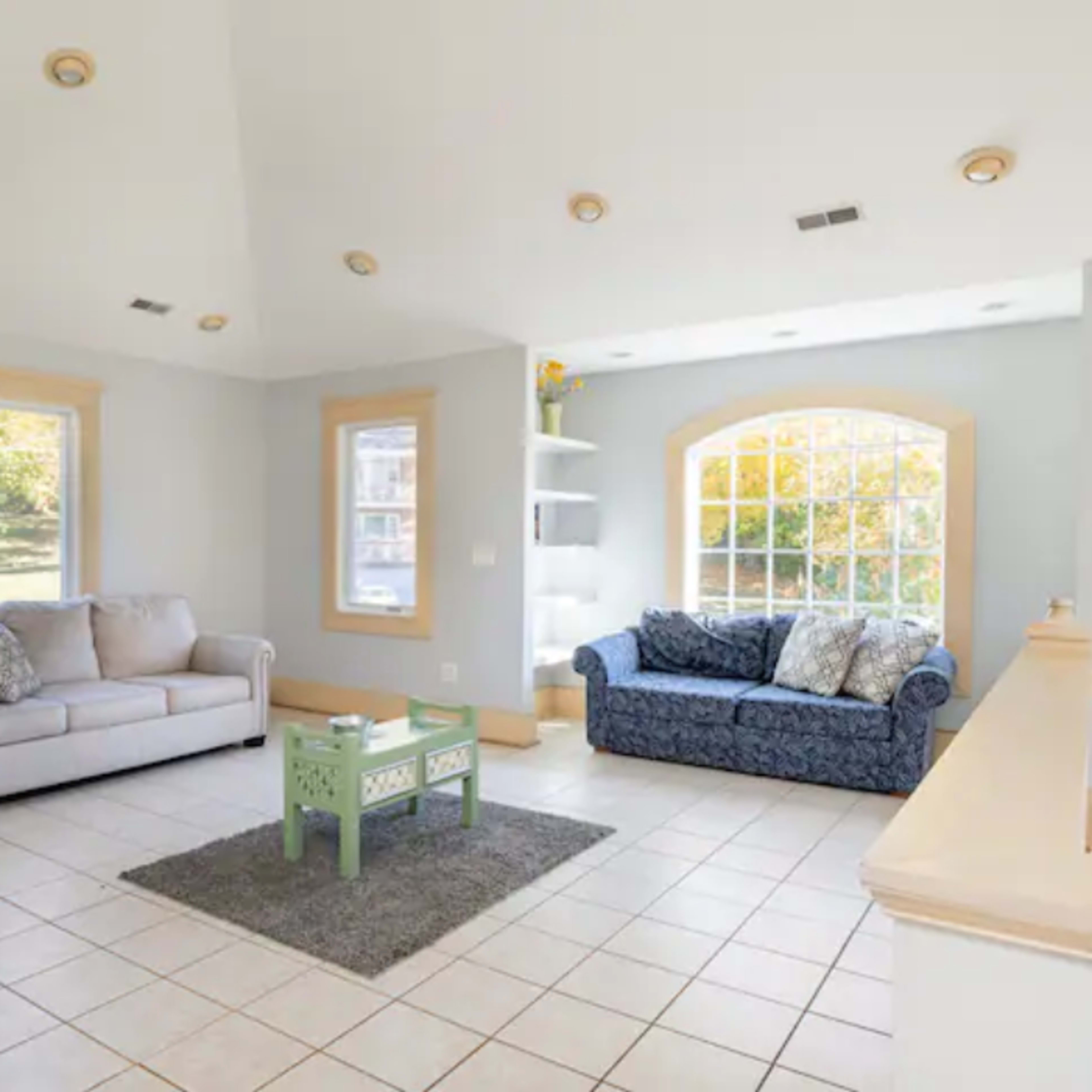 A bright living room features two couches, a green coffee table, and large windows allowing natural light.