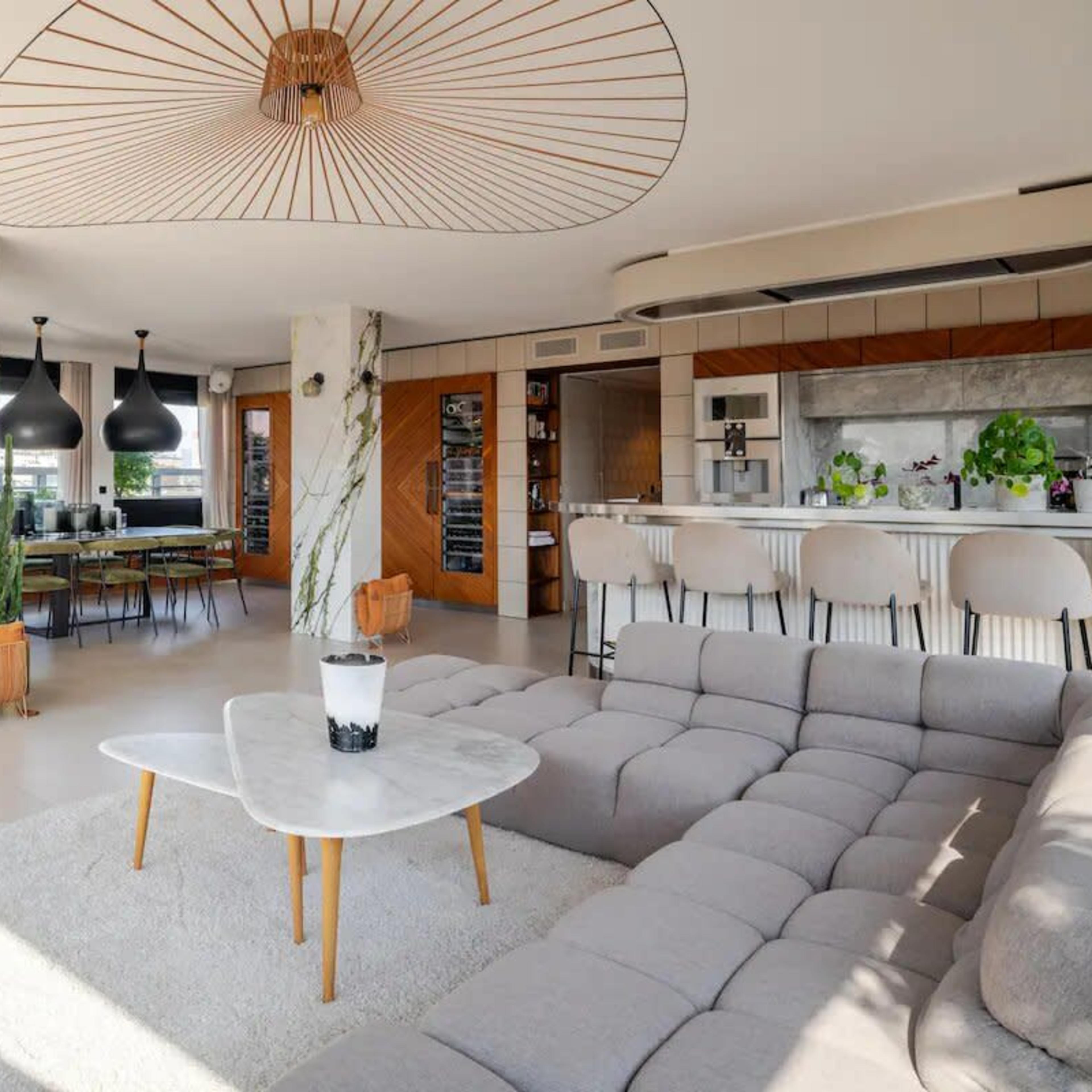 A modern, open-concept living room with a large sectional sofa, a dining area, a piano, and a kitchen featuring an island and green plants for decoration.