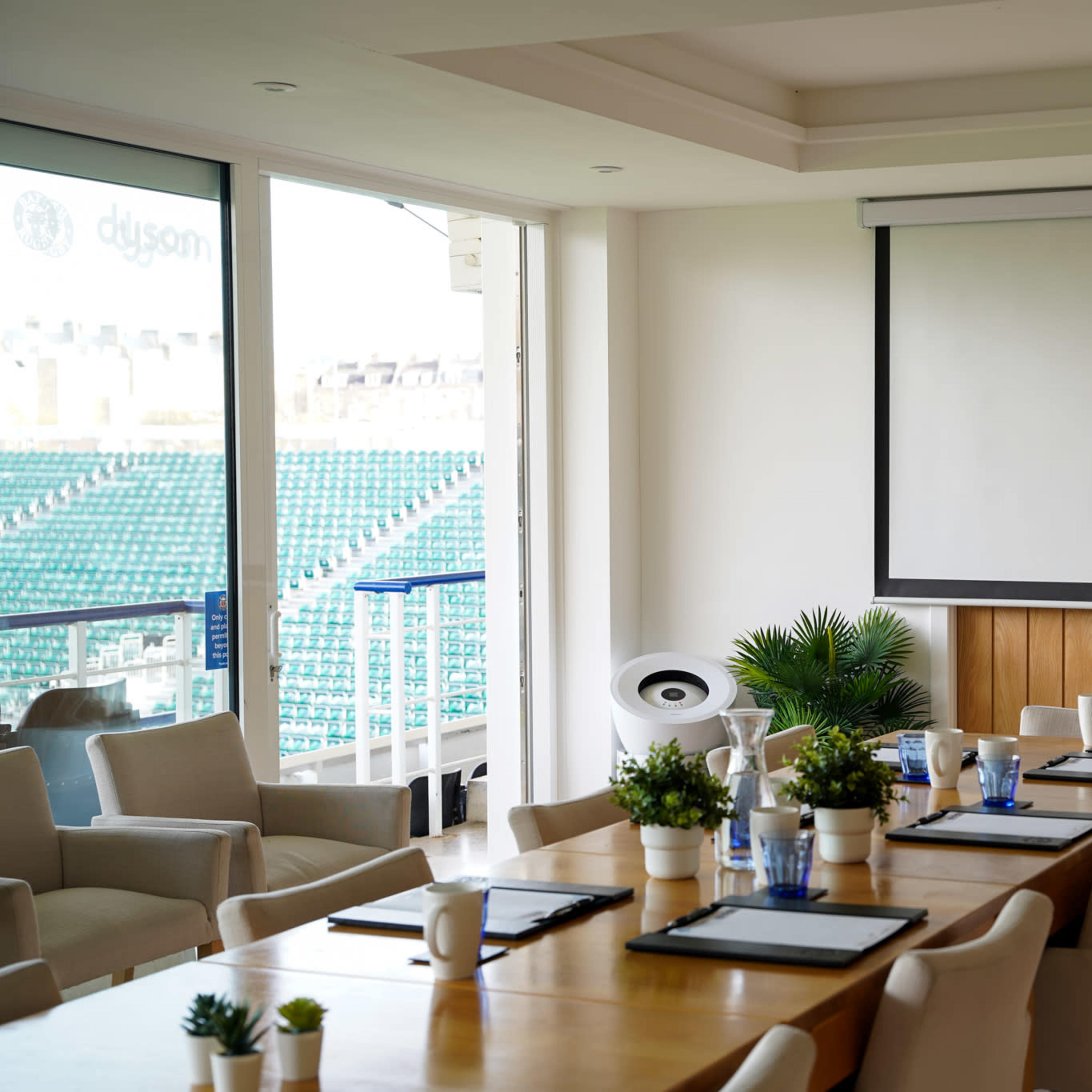 A modern meeting room features a large wooden table set with place settings, a projection screen on the wall, and views of an empty stadium through large windows.