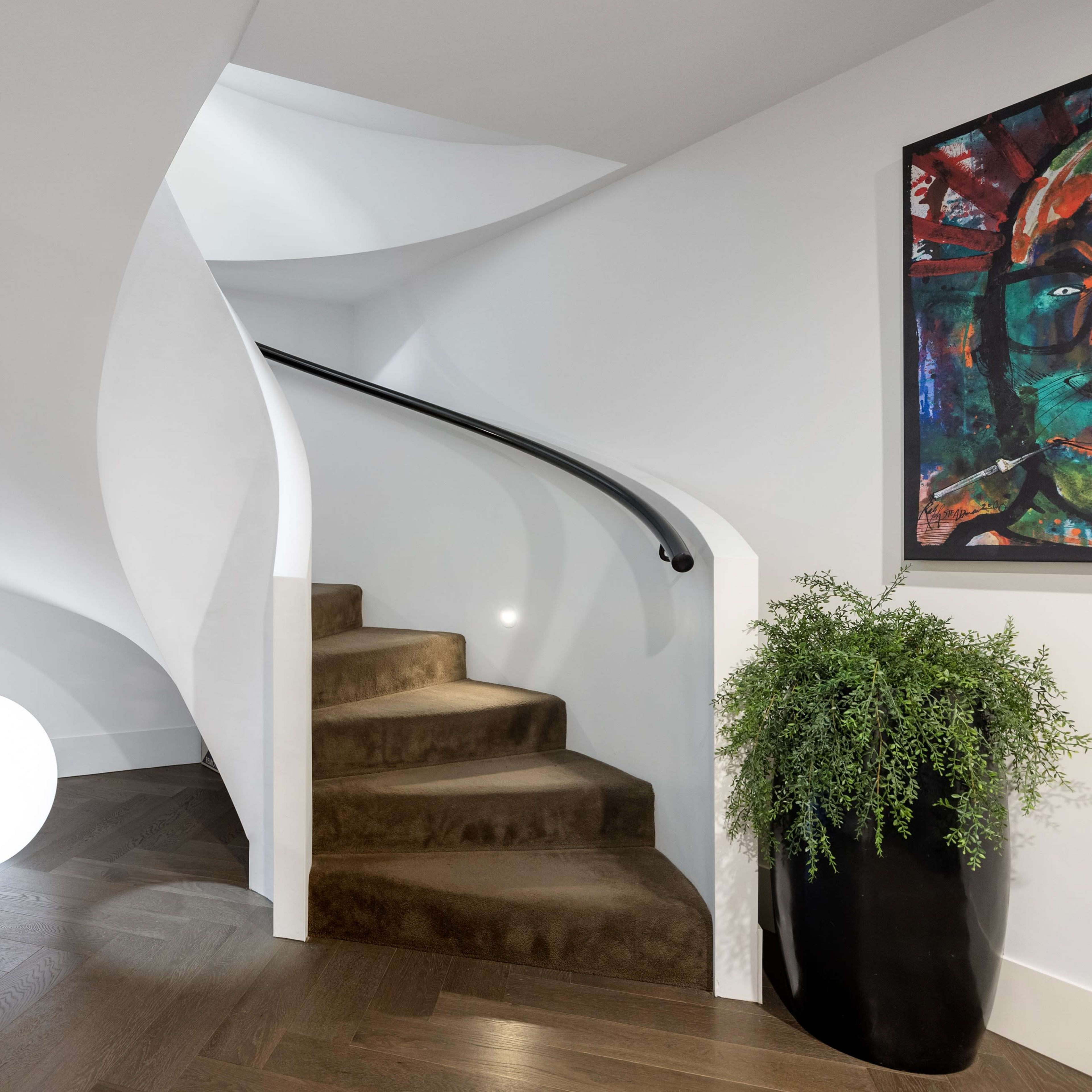 A curved staircase with a dark railing leads up from a light-colored entryway, accompanied by a large round light fixture and potted plants.
