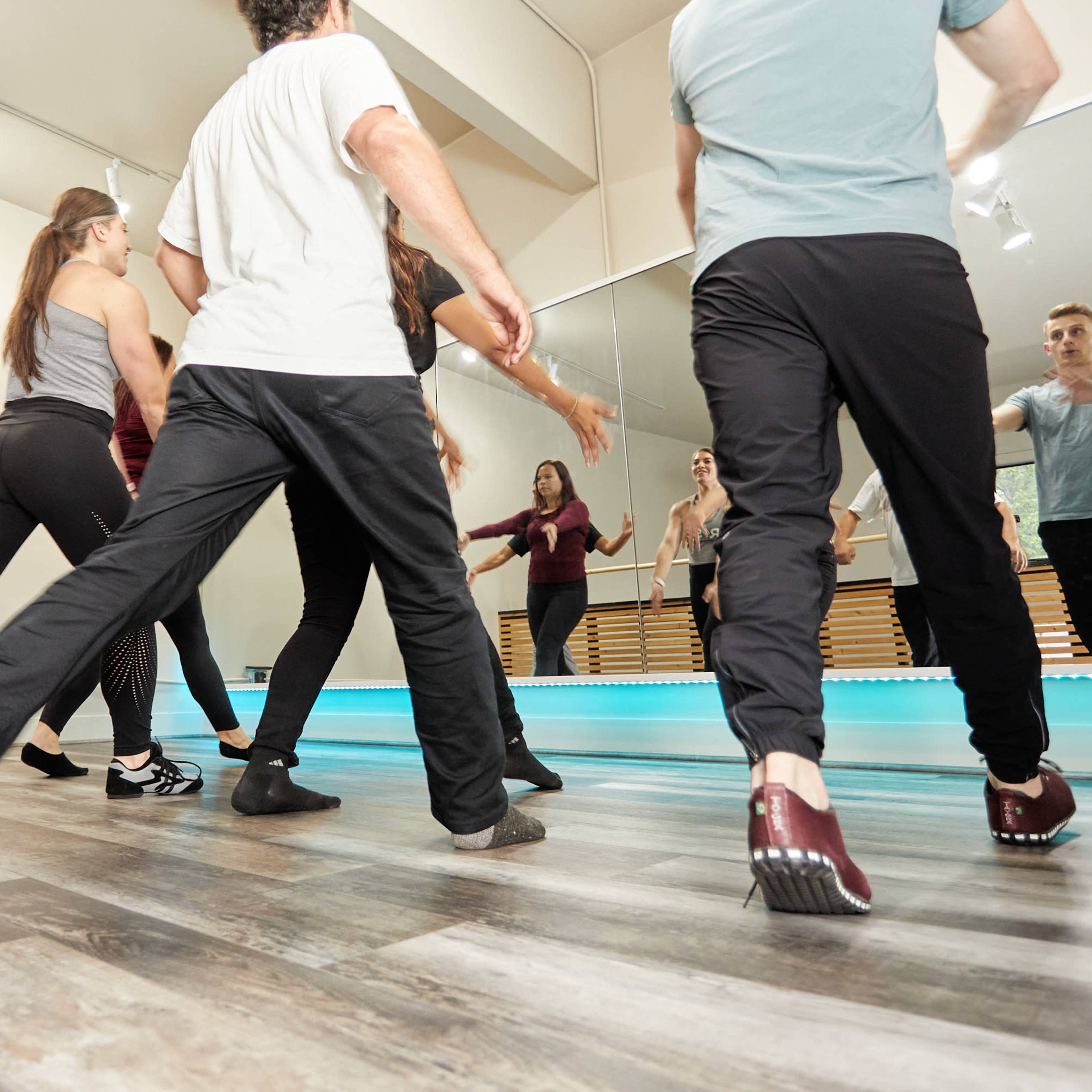 A group of dancers practice their movements in a well-lit studio with mirrors and a wooden floor.