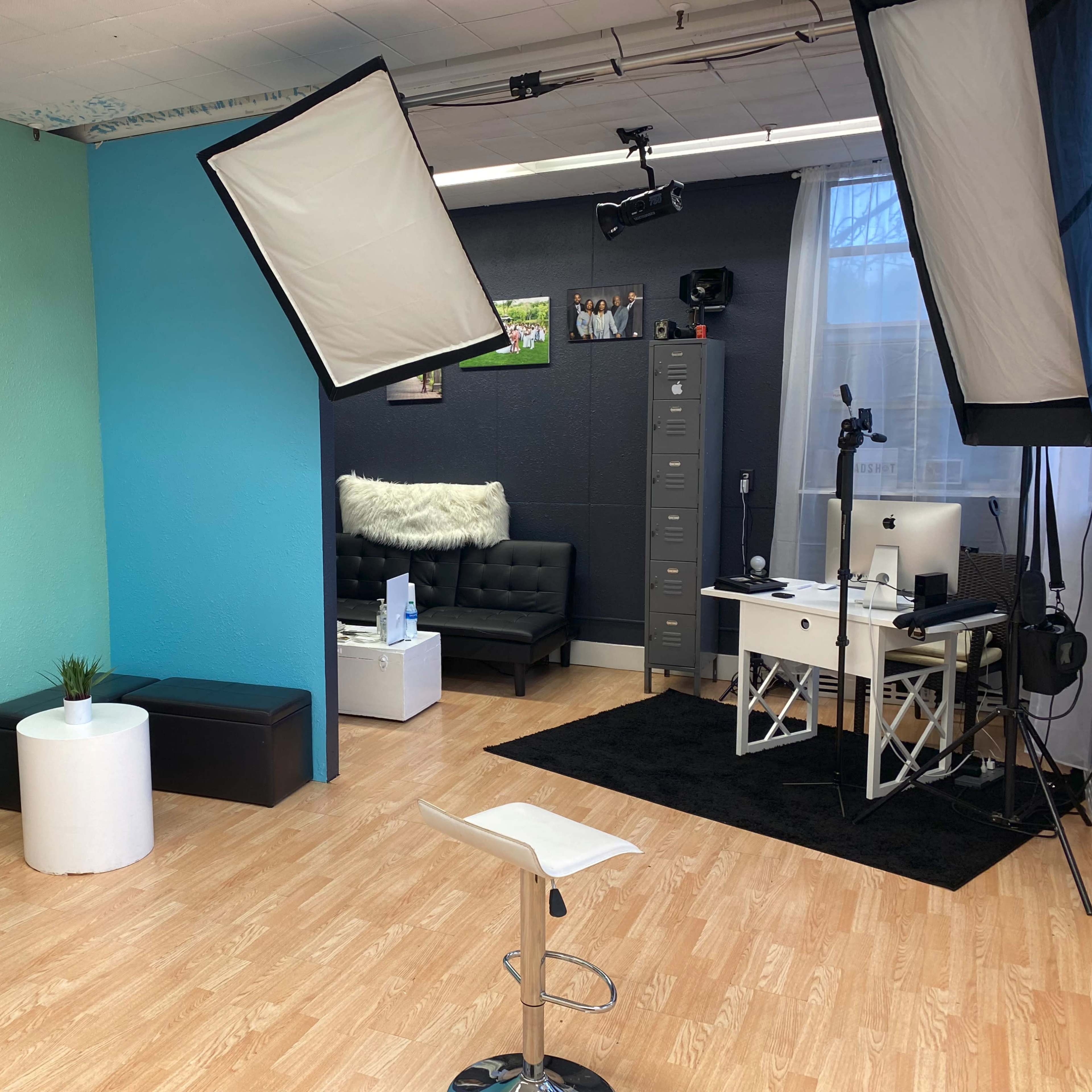 A spacious studio with different colored wall sections, a black couch, a white desk with a computer, and lighting equipment set up for a photoshoot.