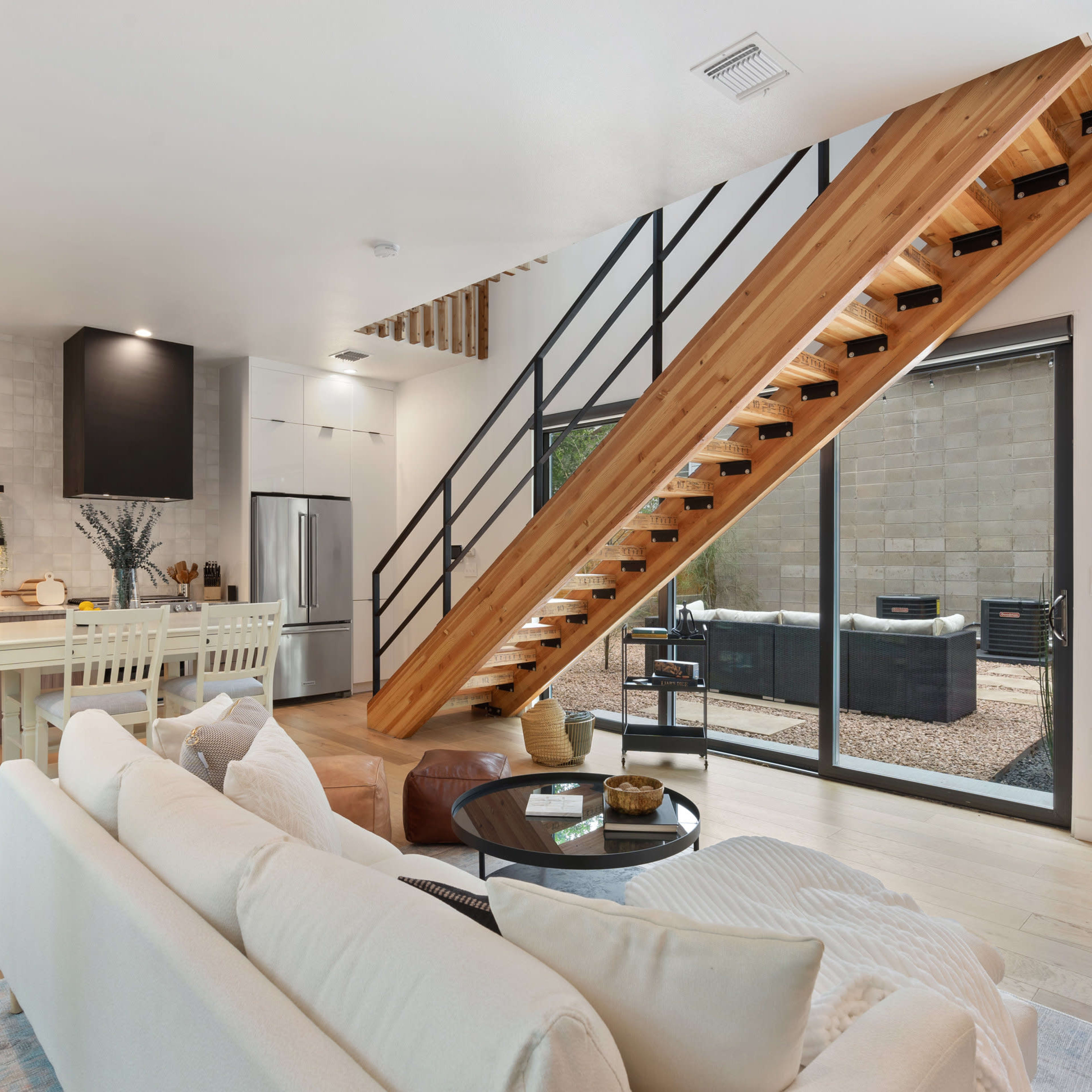 A modern interior features a living area with a white sofa, a kitchen with stainless steel appliances, and a wooden staircase leading to a second floor.