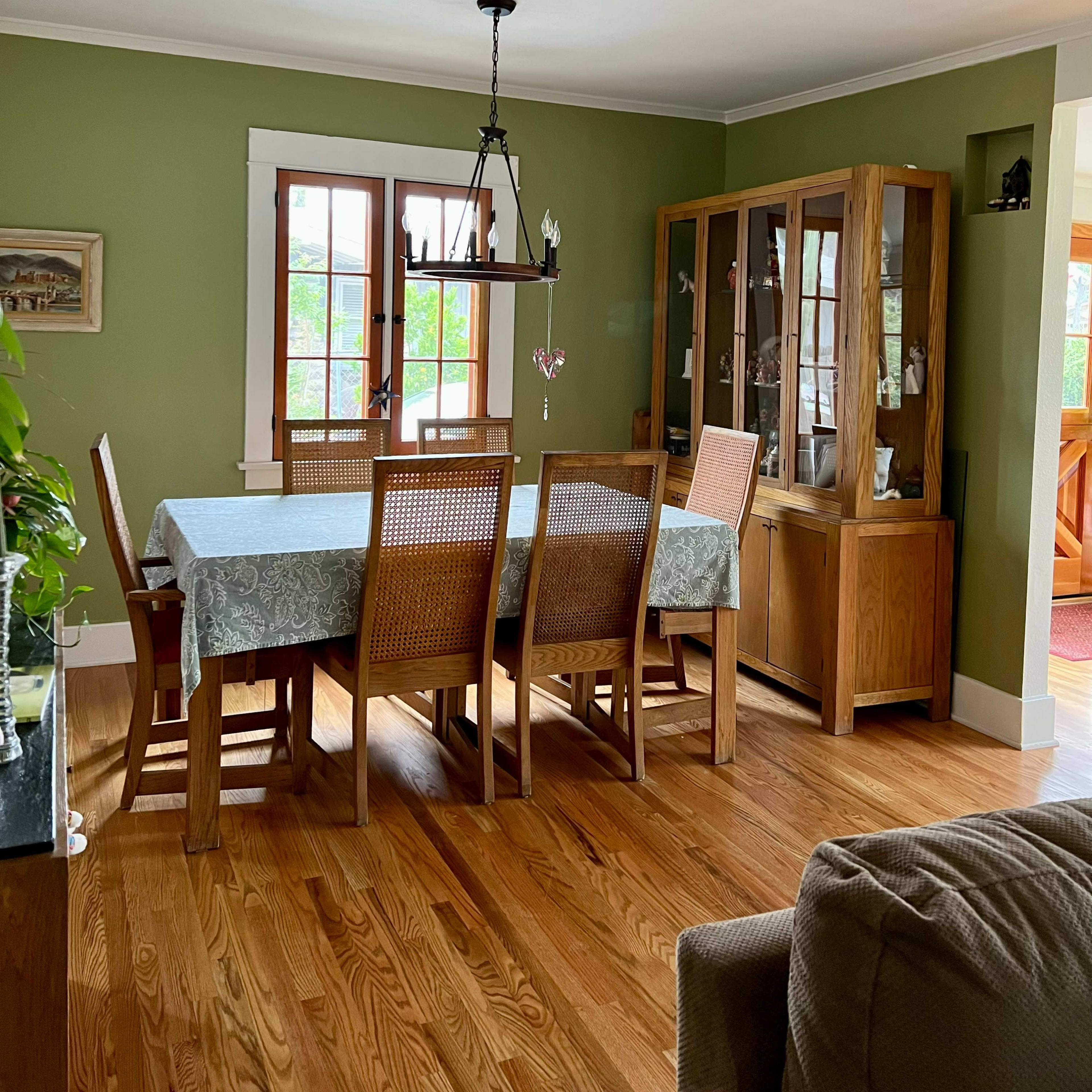 A dining room features a large wooden table surrounded by chairs, a glass-fronted cabinet, and green walls with natural light streaming in through windows.