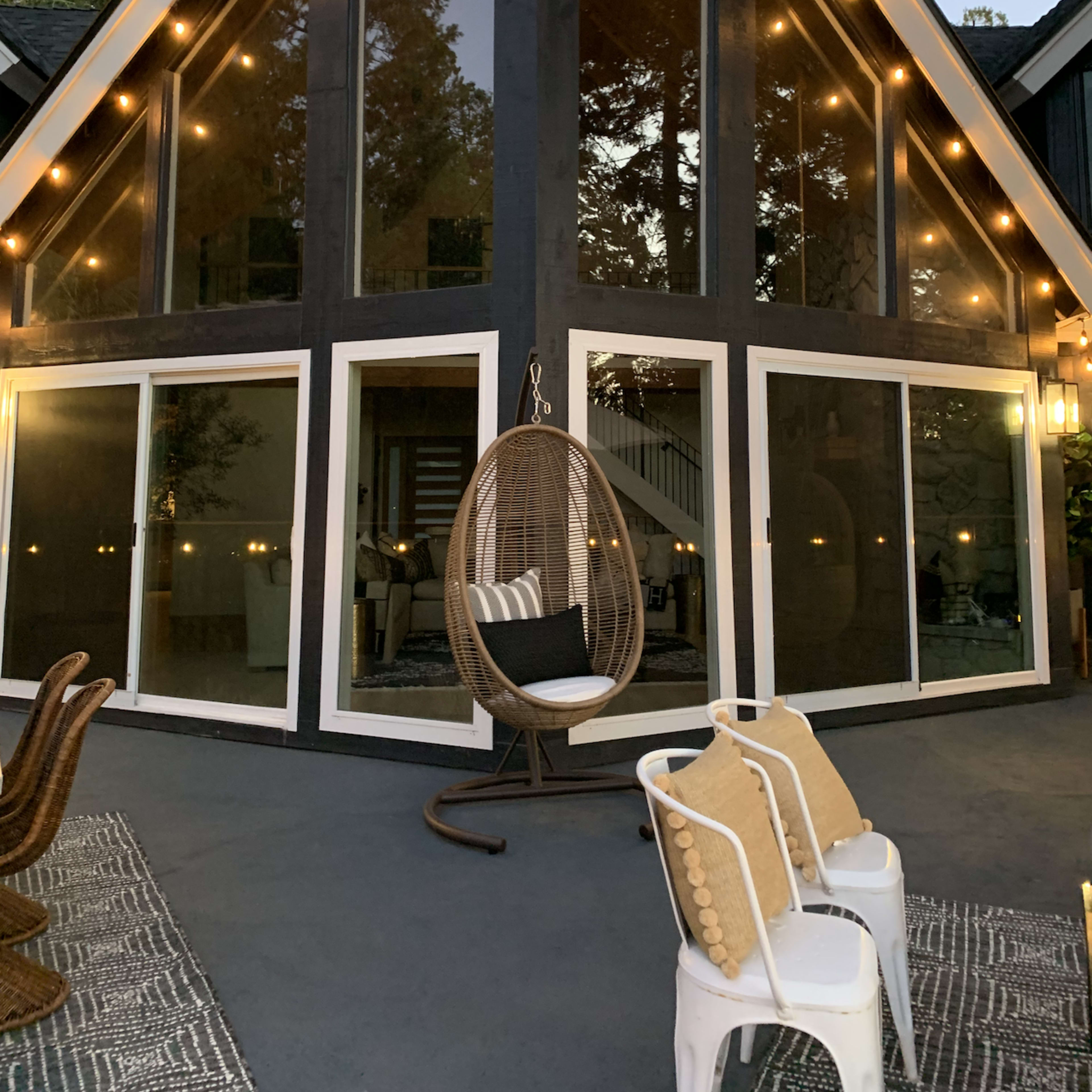 A modern A-frame house with large glass windows, surrounded by trees and illuminated by string lights.
