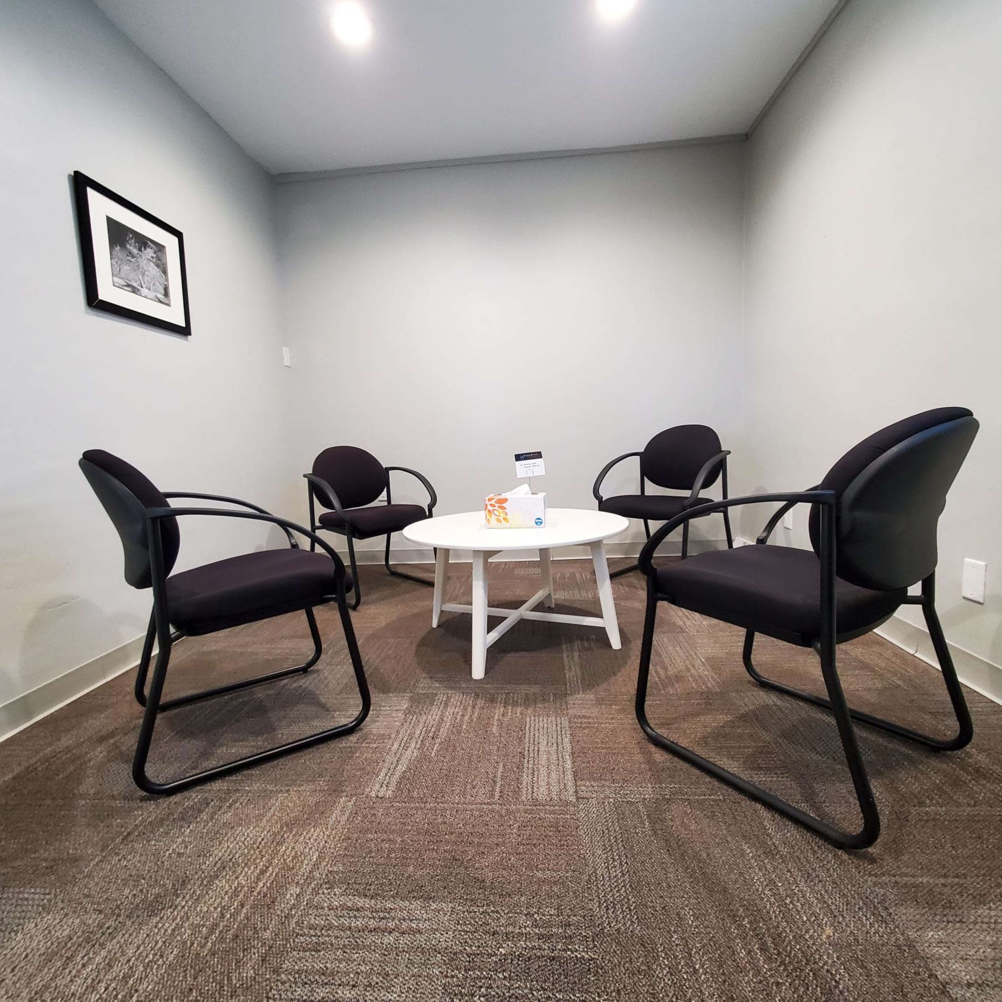 A small meeting room with four black chairs arranged around a white table, set against a gray wall.
