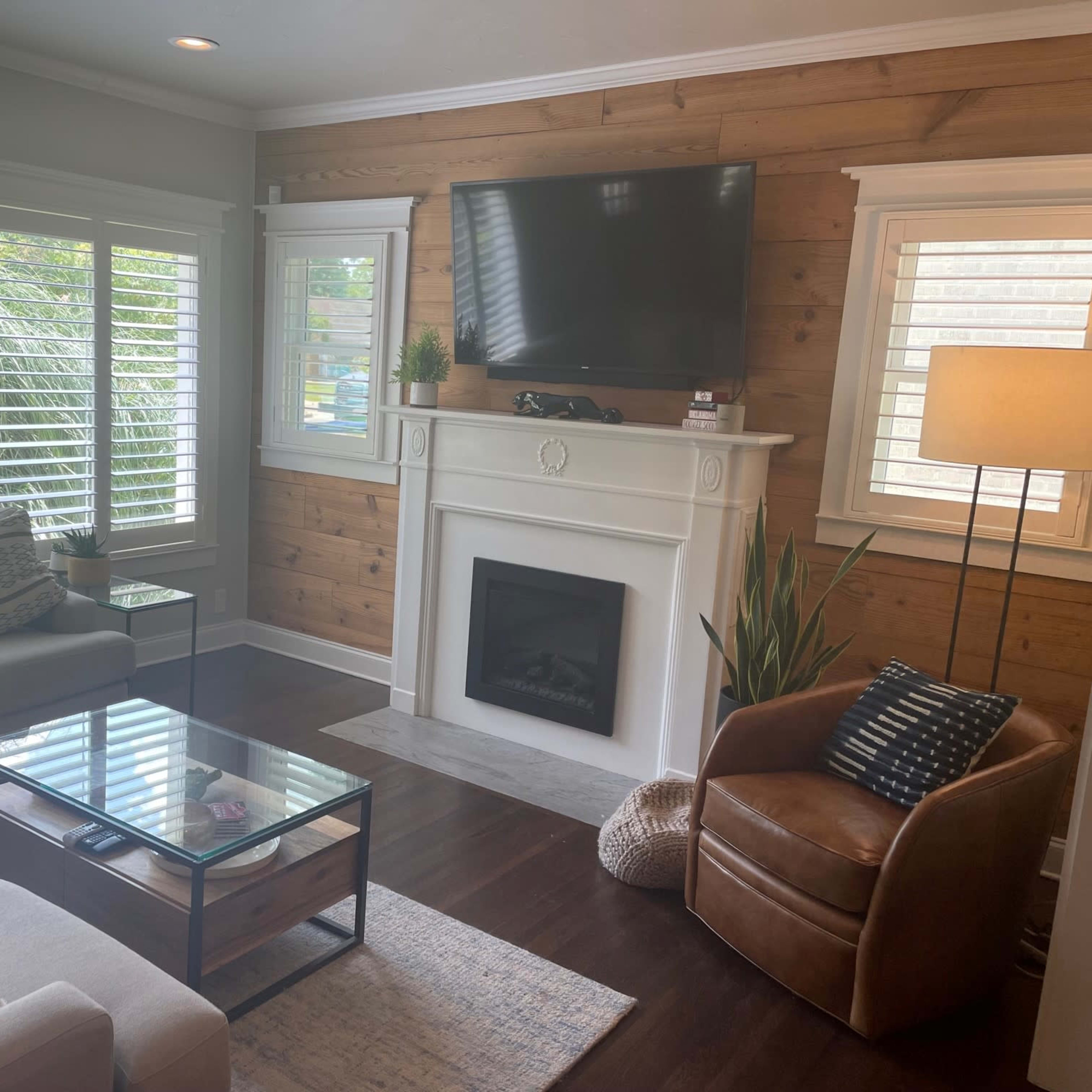 A living room features a sectional sofa, a glass coffee table, a fireplace with a white mantel, and large windows with shutters letting in natural light.