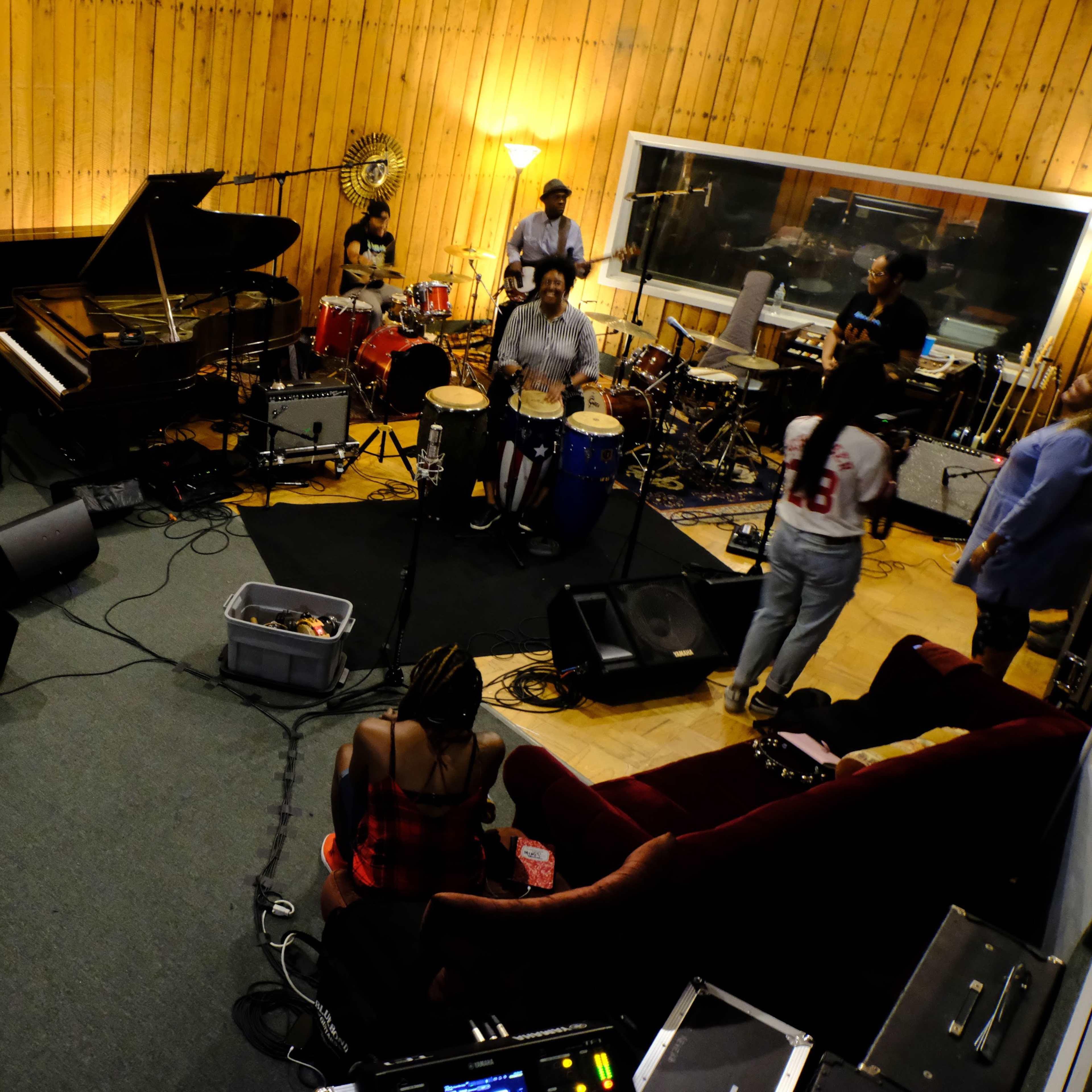 A group of musicians performs in a recording studio while an audience sits nearby, observing the session.
