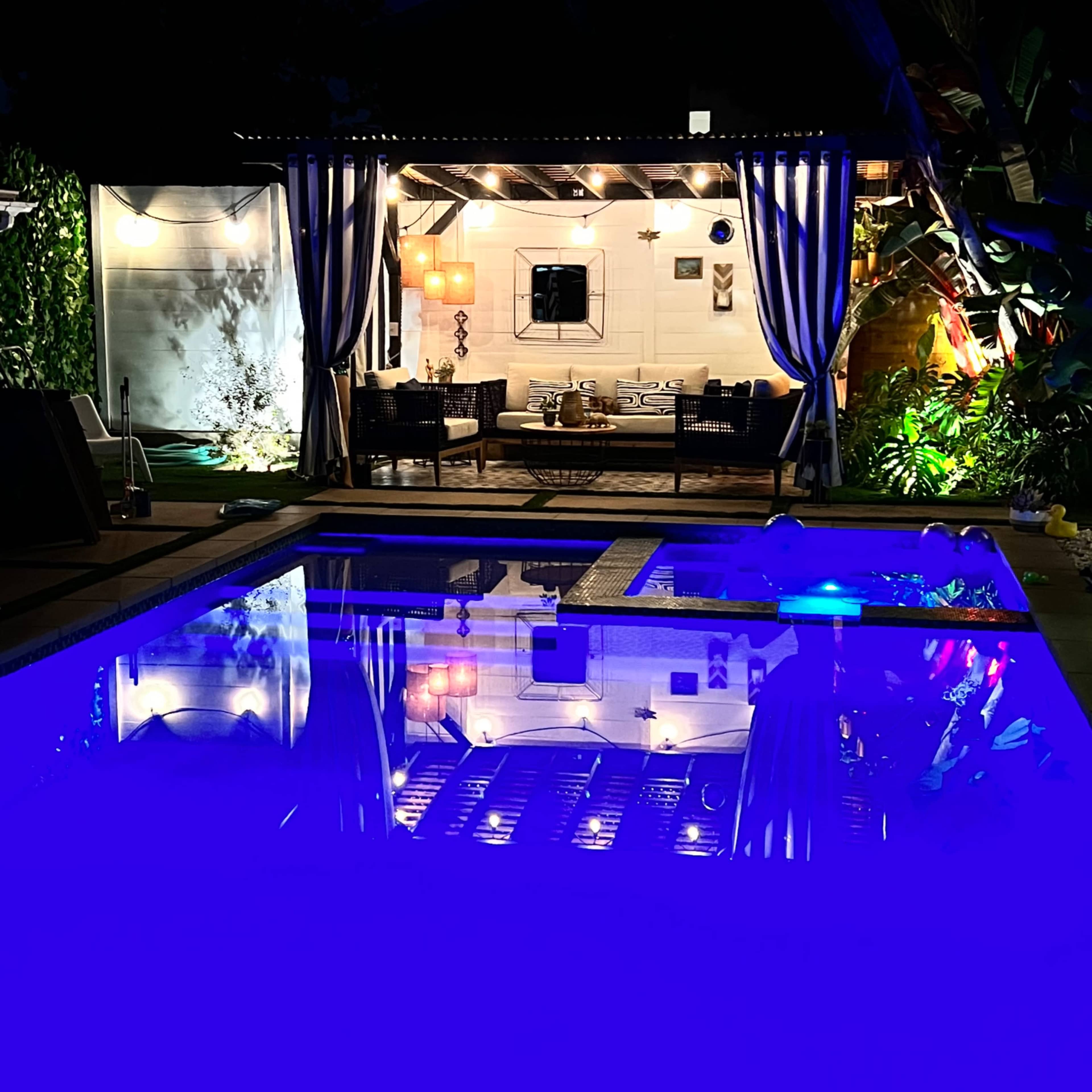 A well-lit outdoor lounge area with seating is reflected in a pool at night, surrounded by greenery and illuminated by ambient lighting.