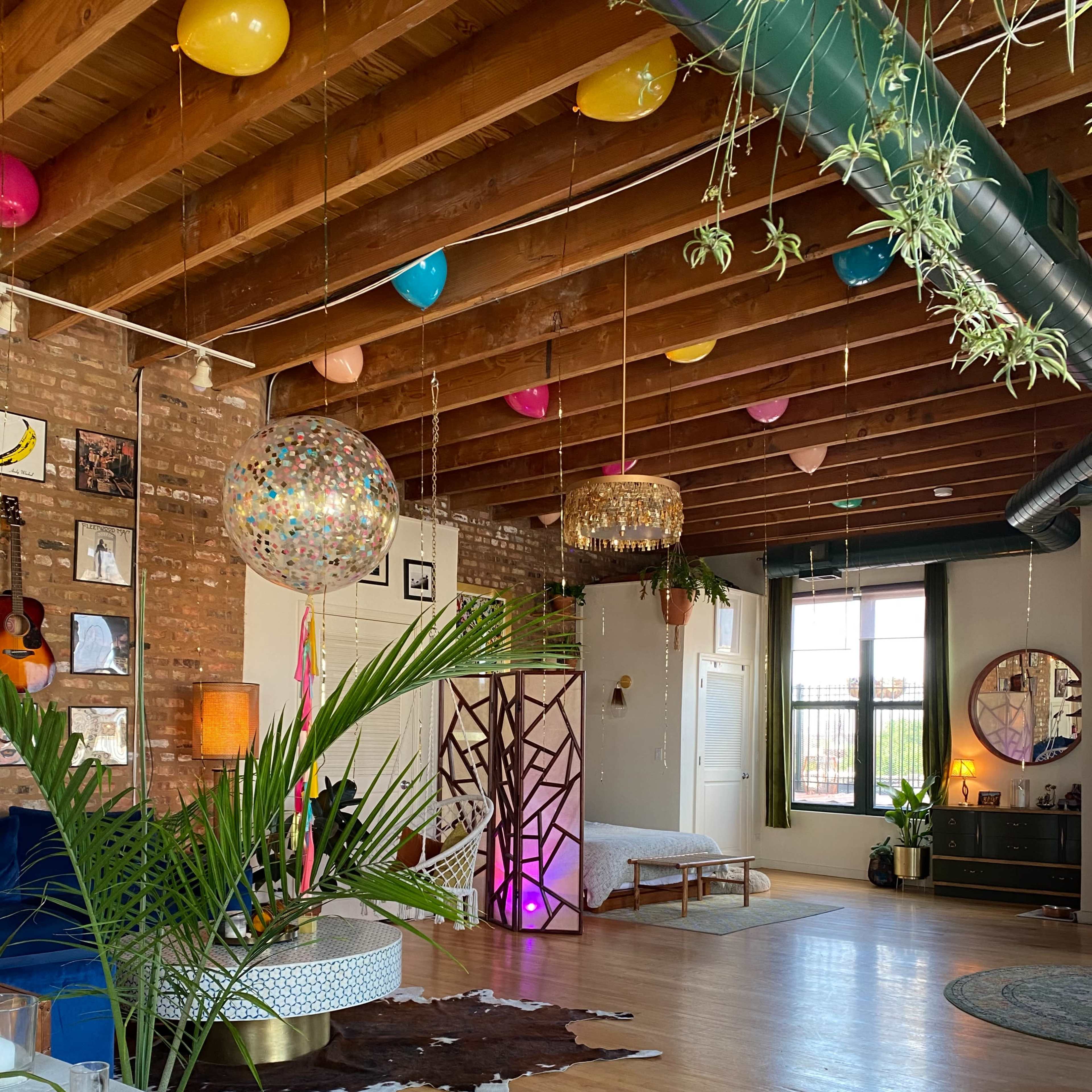 A spacious room features exposed wooden beams, colorful balloons hanging from the ceiling, and a mix of decorative plants and furniture.