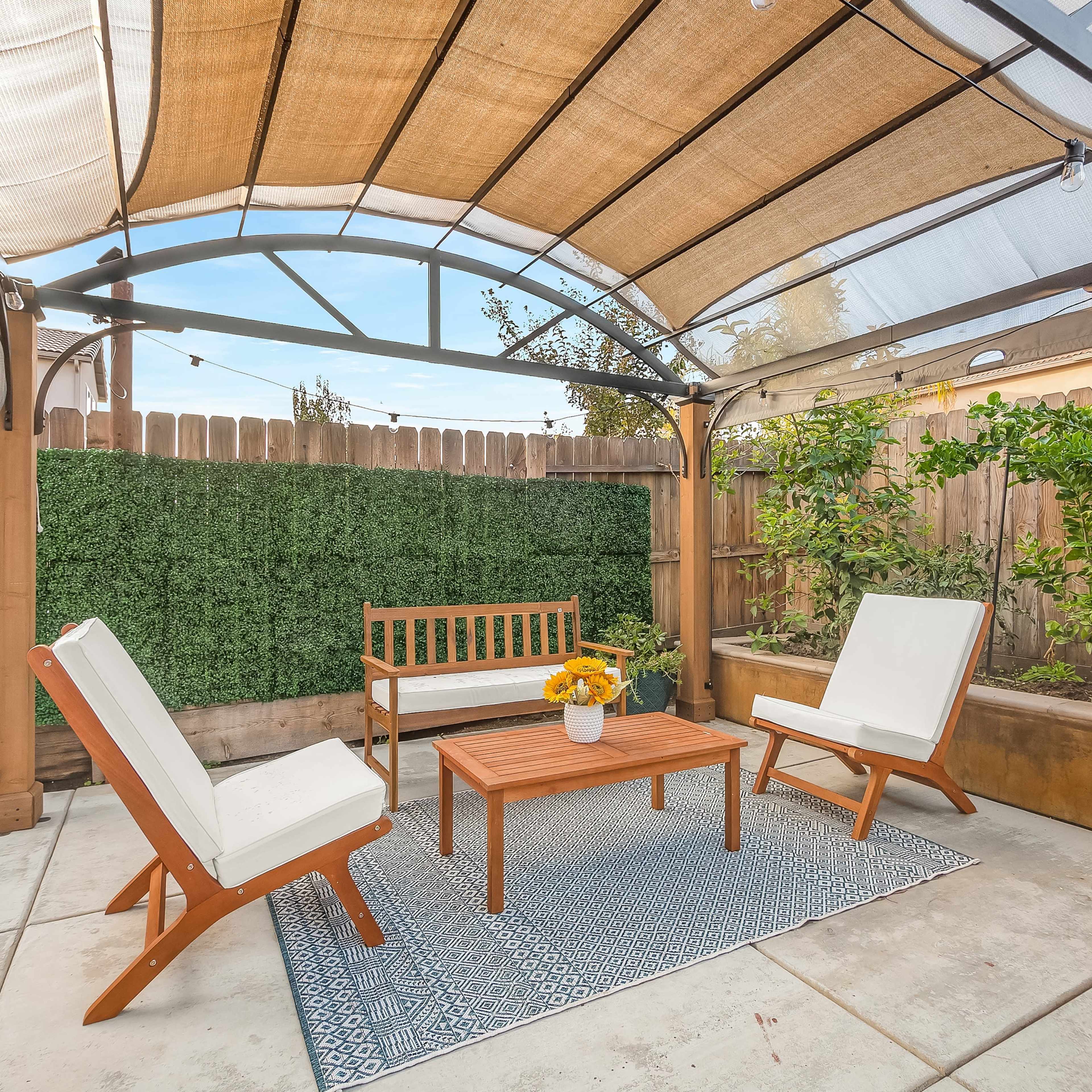A covered outdoor seating area features two white chairs, a table, and a bench surrounded by green hedges.