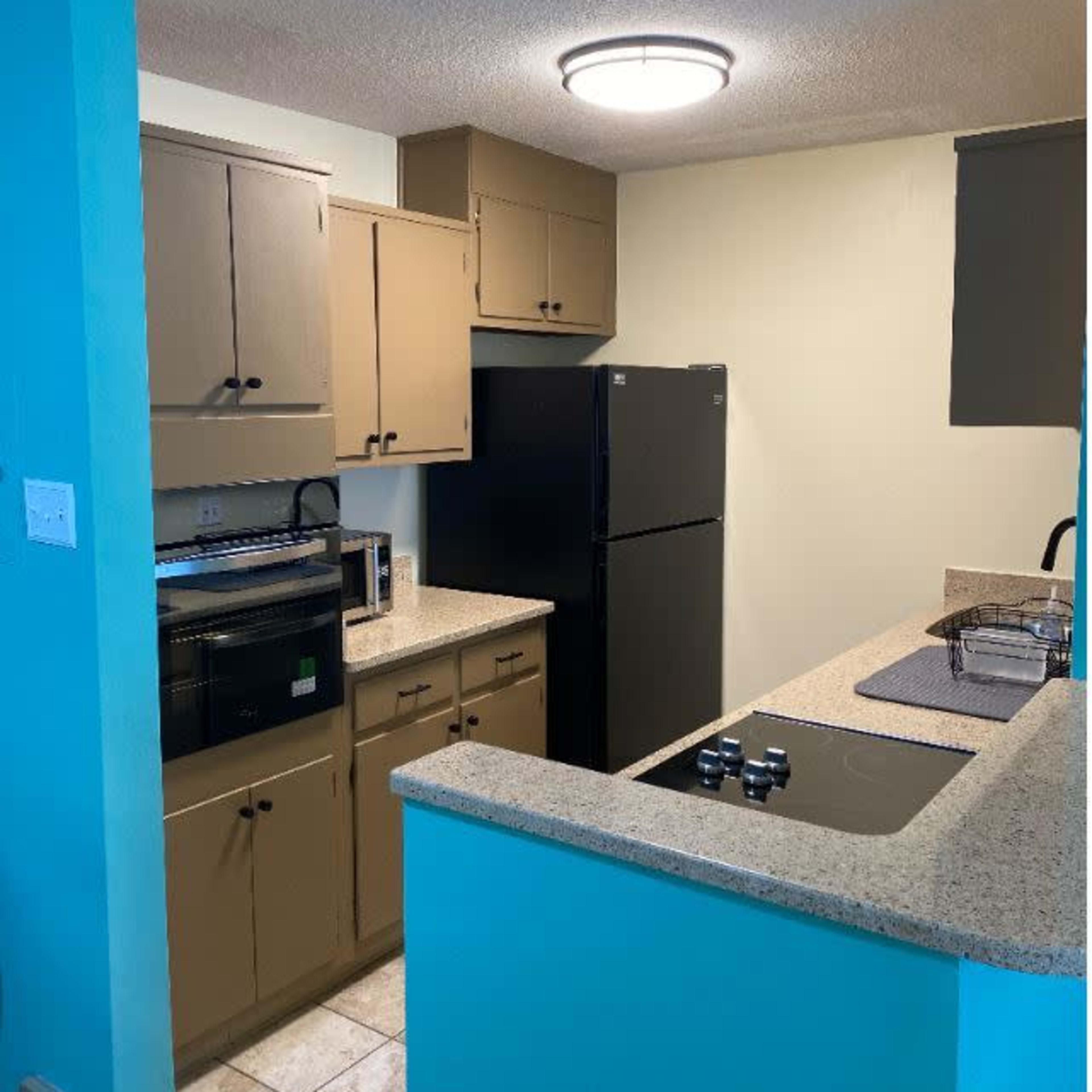 A kitchen featuring beige cabinets, a black refrigerator, a stove with a cooktop, and light-colored countertops against a blue wall.