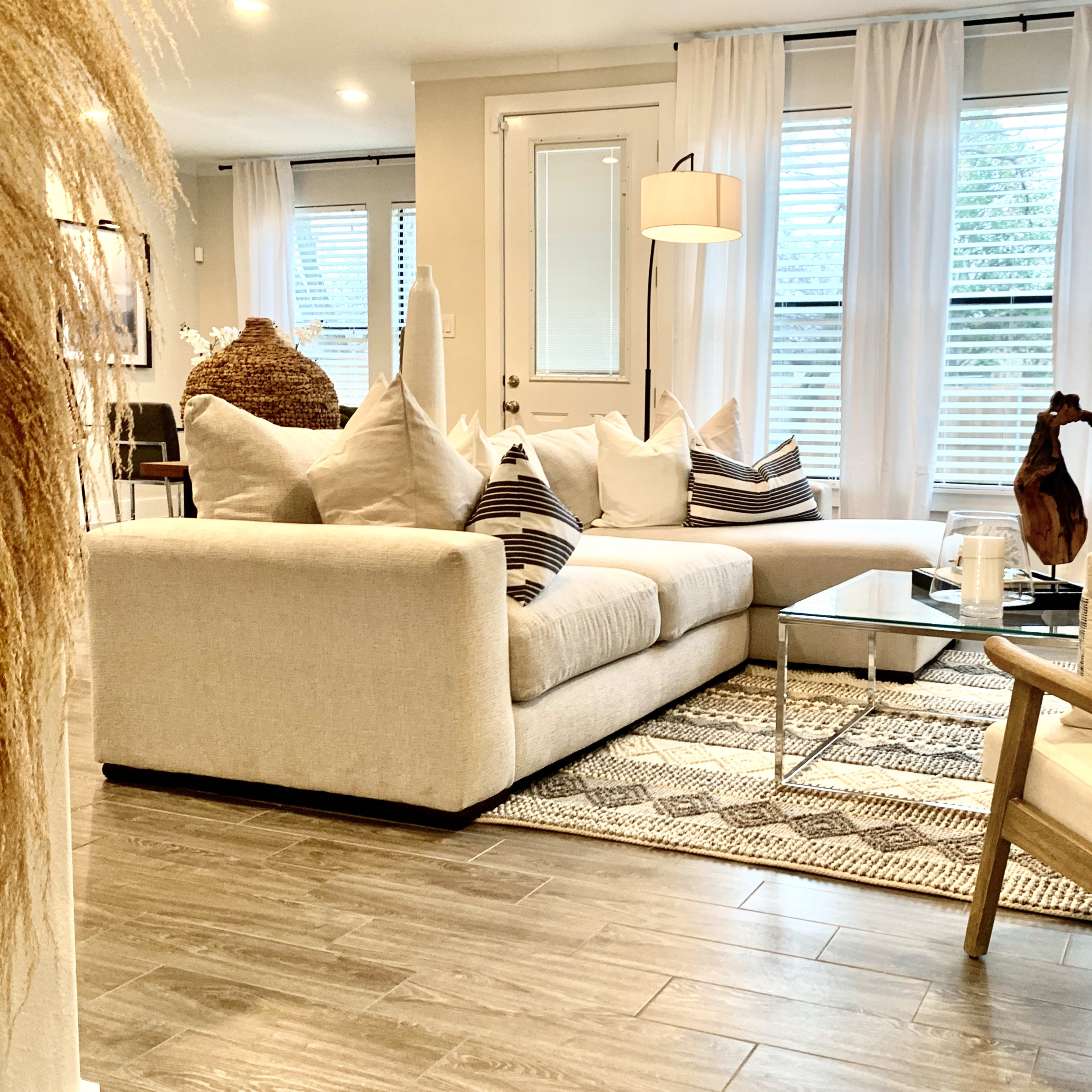 A modern living room features a light-colored sectional sofa, a glass coffee table, and large windows adorned with sheer curtains.