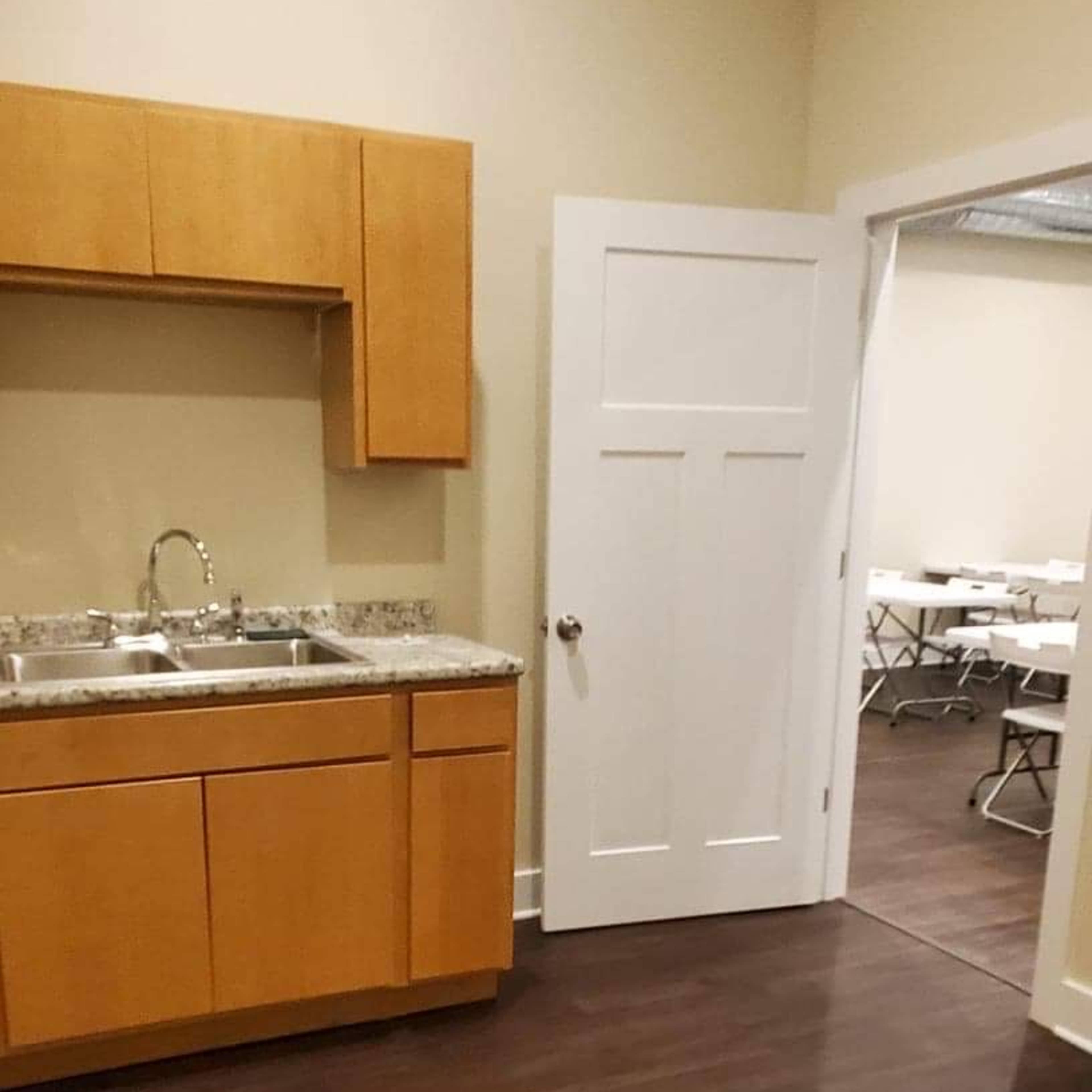 A simple kitchen area with wooden cabinets, a countertop, and a sink, adjacent to a doorway leading to another room with folding tables.