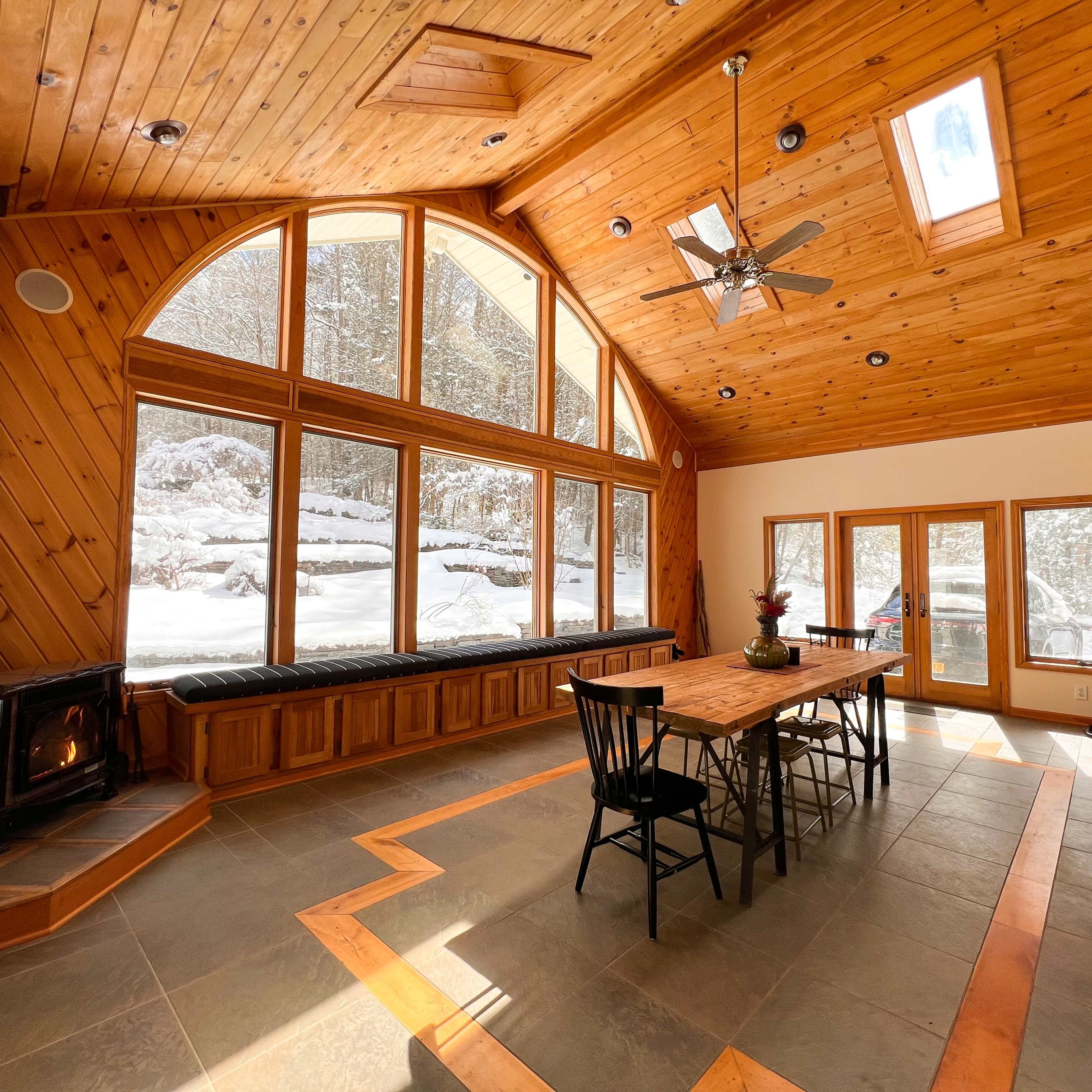 A spacious wooden interior with large windows overlooking a snowy landscape, featuring a dining table at the center and a wood stove in the corner.