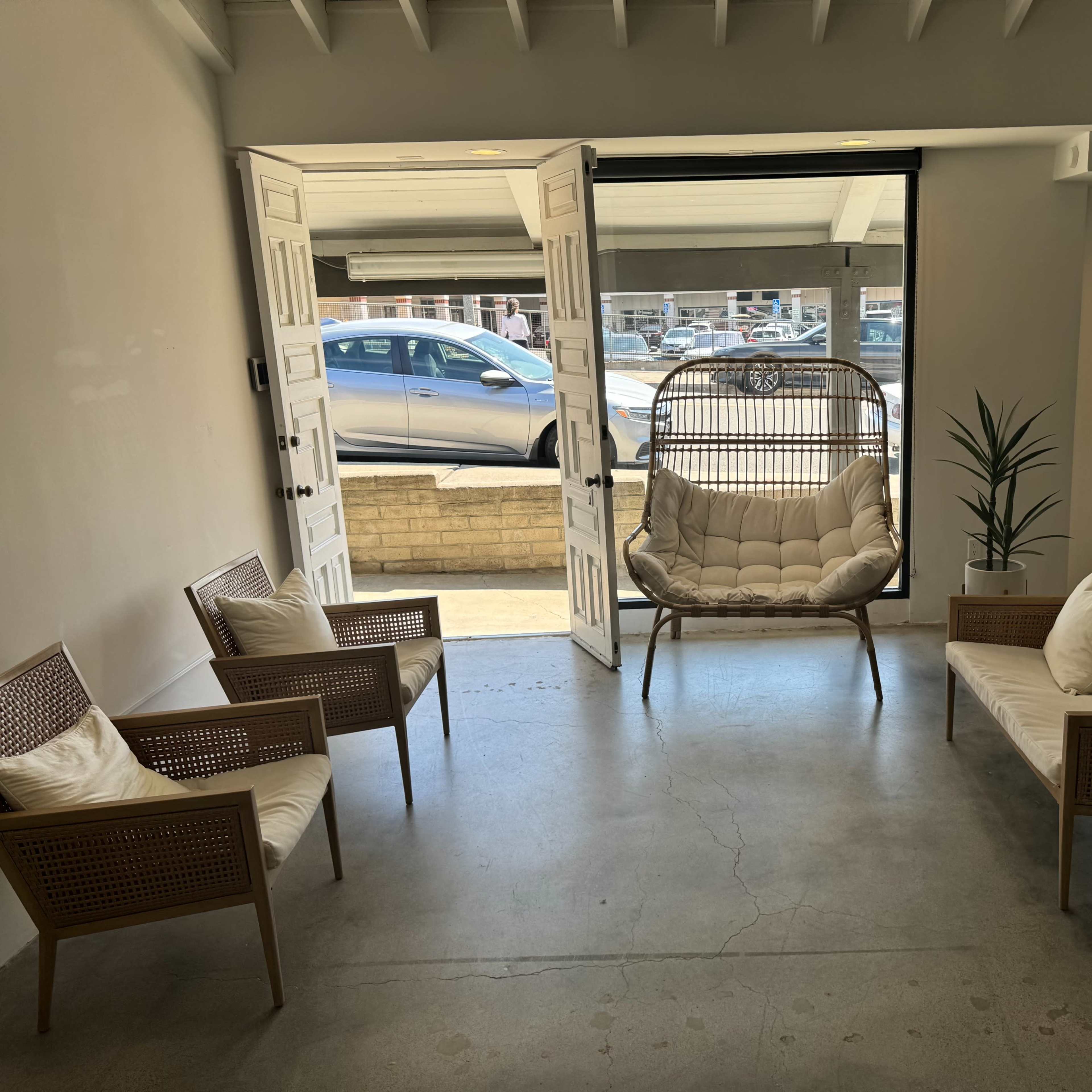 A modern lounge area with several chairs and a hanging chair, facing open double doors that lead to a parking lot.