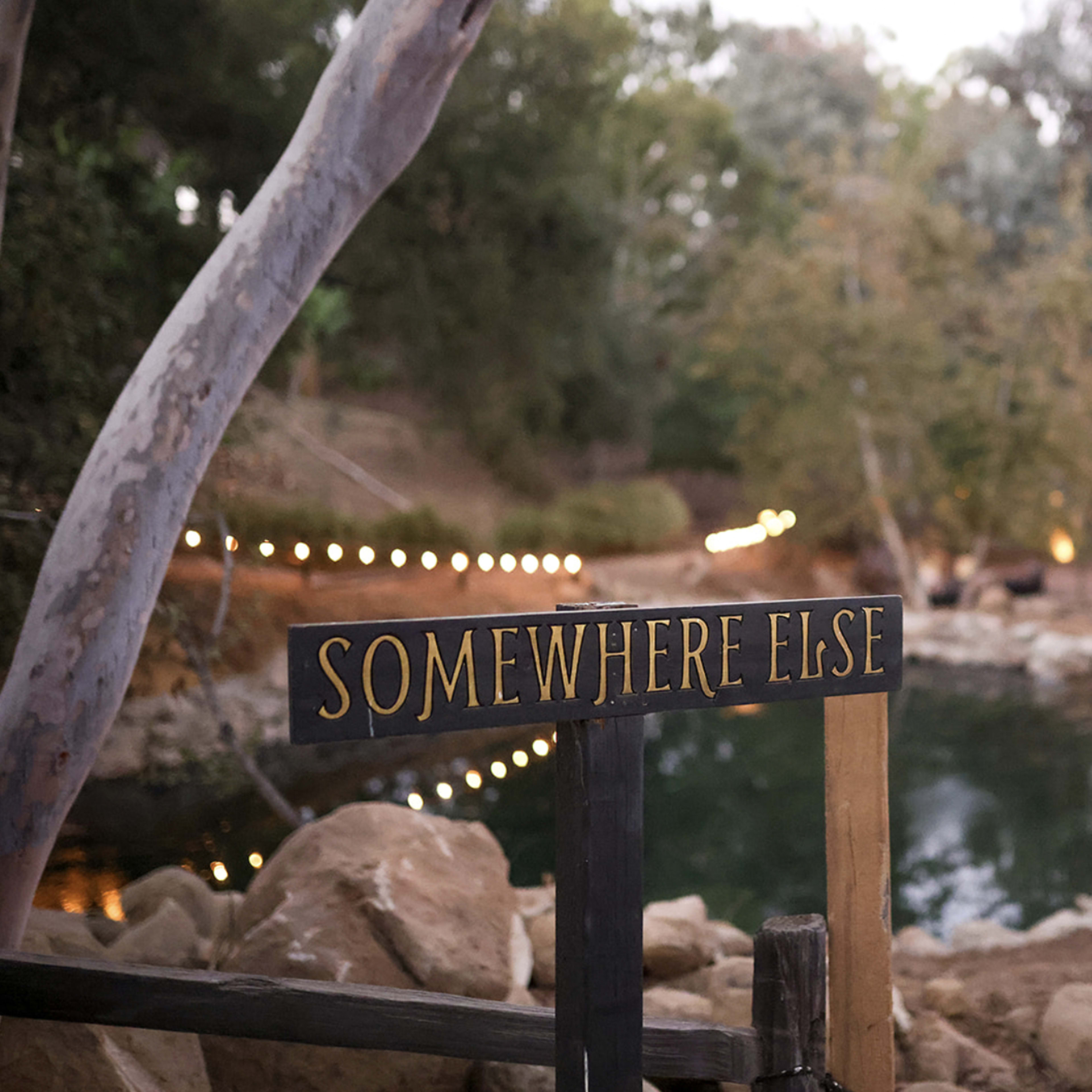 A wooden sign reading "Somewhere Else" stands near a small pond surrounded by trees and a pathway illuminated by string lights.