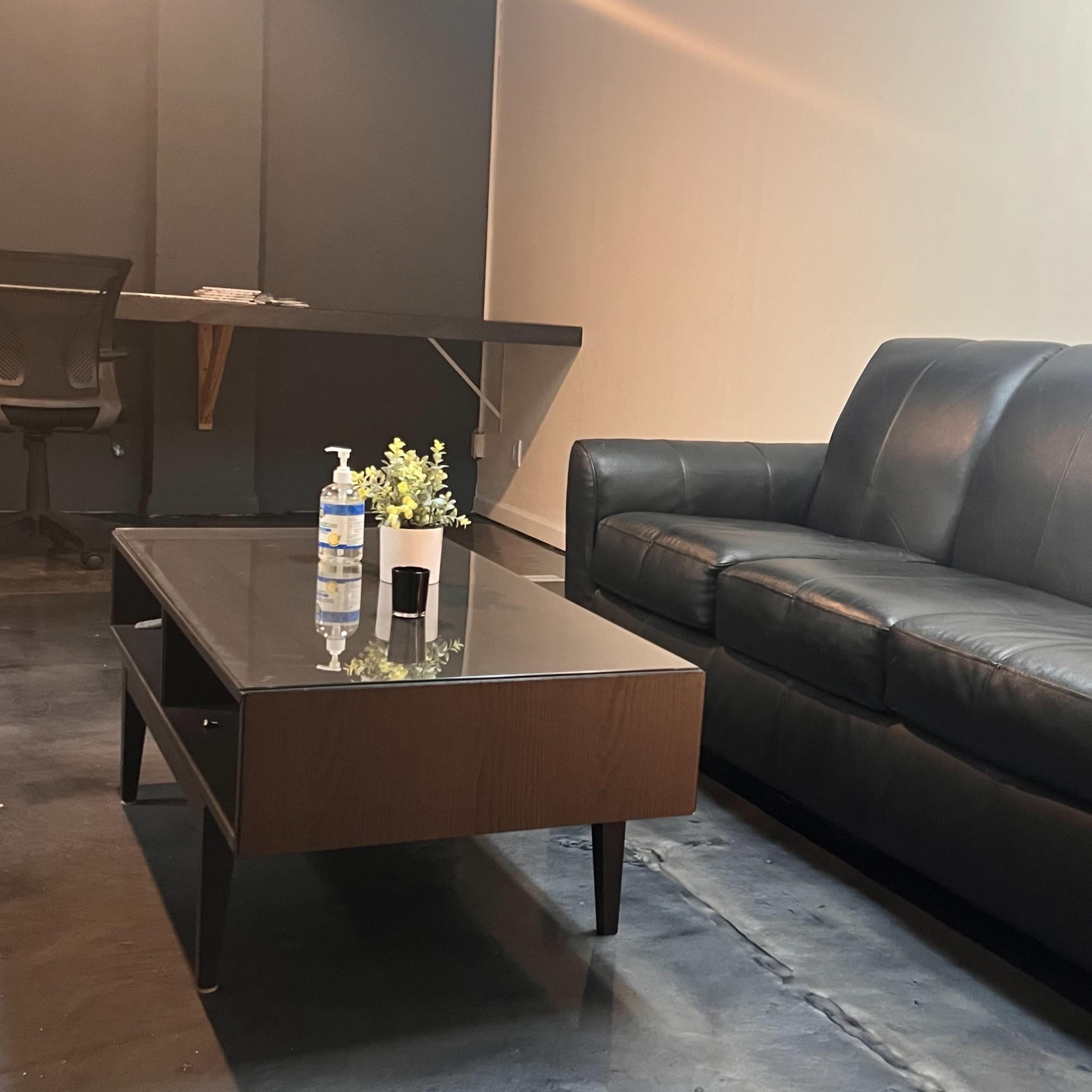 A minimalist room with a black leather couch, a coffee table, and a desk with a chair against a plain wall.
