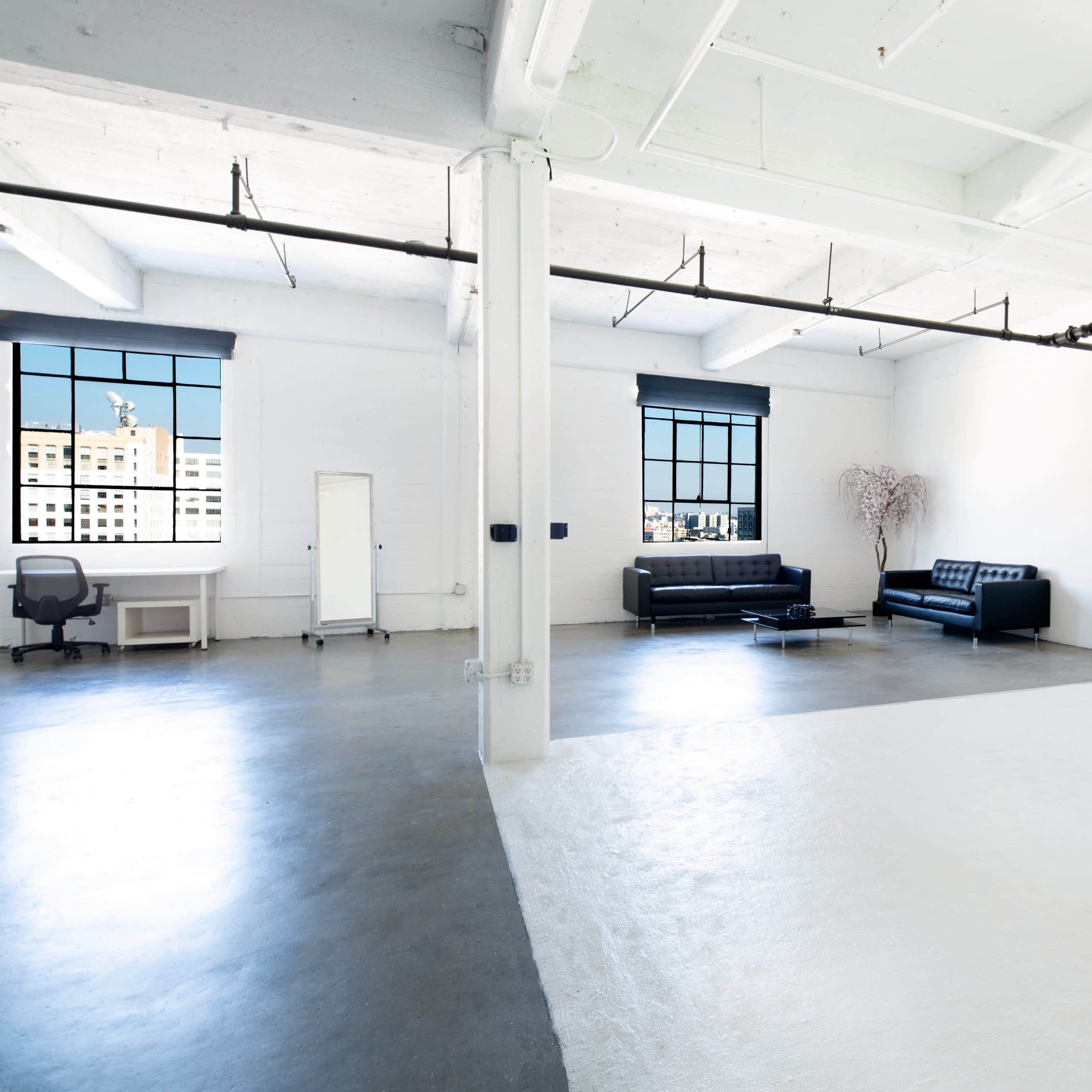 A spacious, minimalist room featuring large windows, a black sofa, a desk with a chair, and a white pedestal on a divided floor.