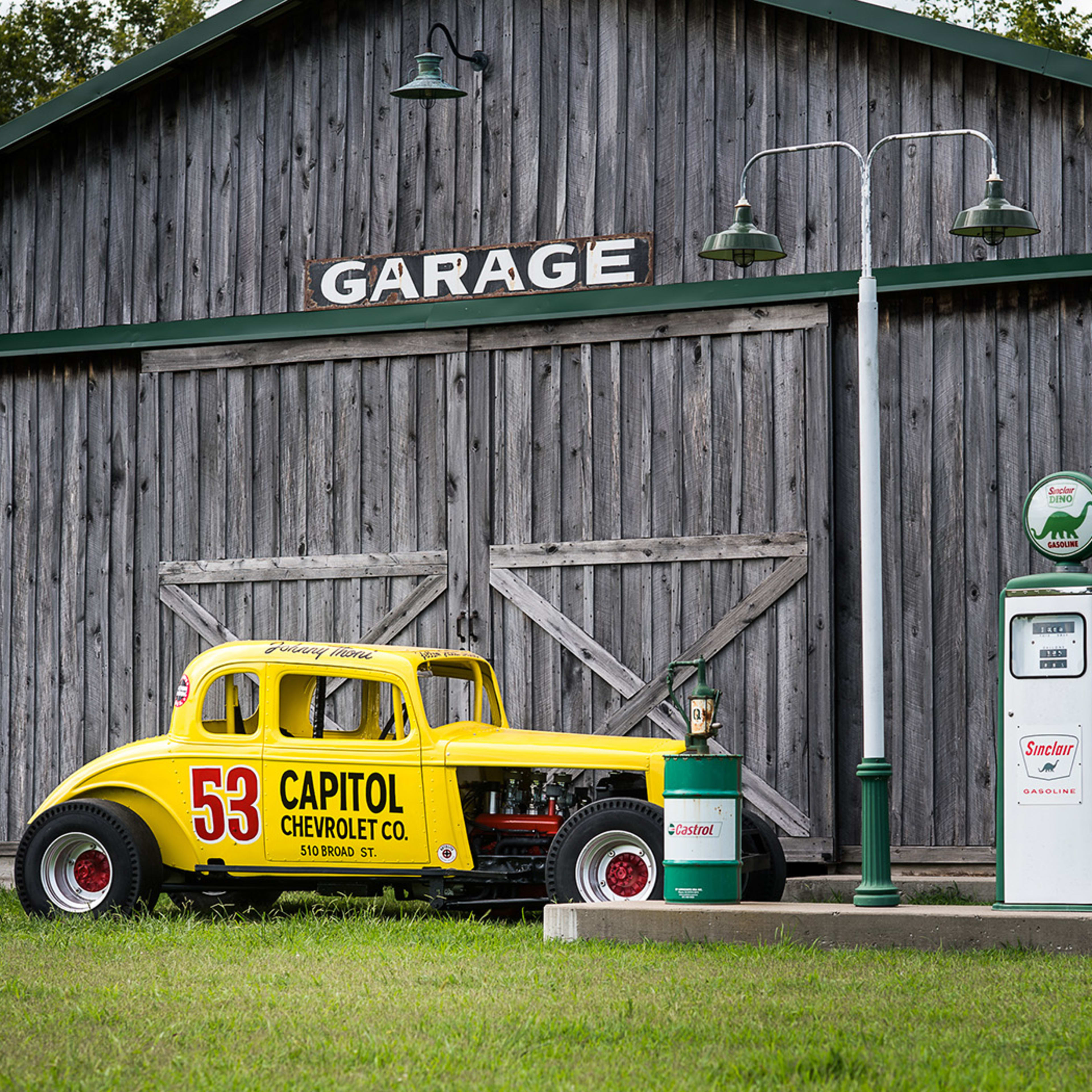 A yellow vintage race car with the number 53 is parked beside a green and white gas pump in front of a rustic wooden garage.
