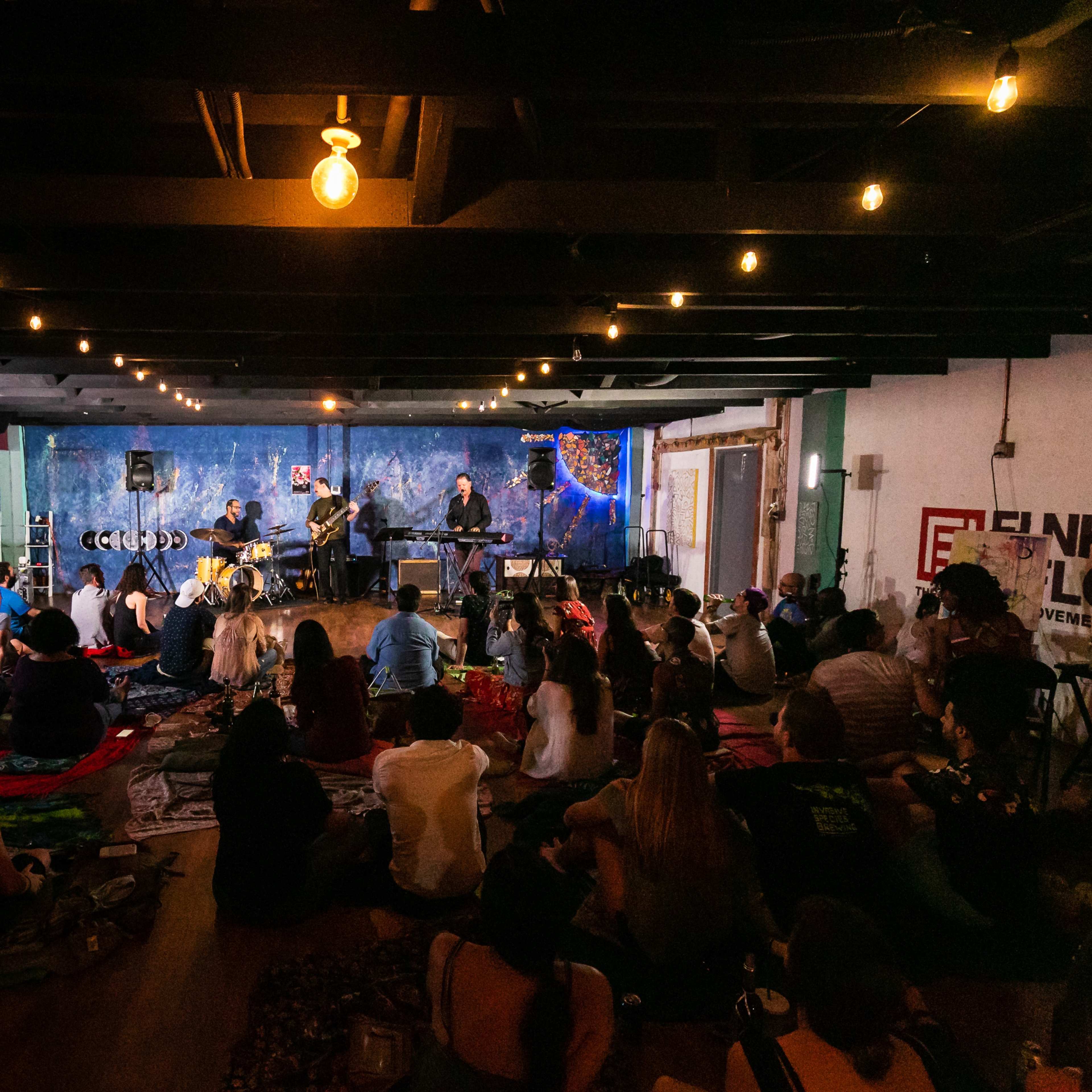 A live music performance is taking place in a dimly lit room with an audience seated on the floor.