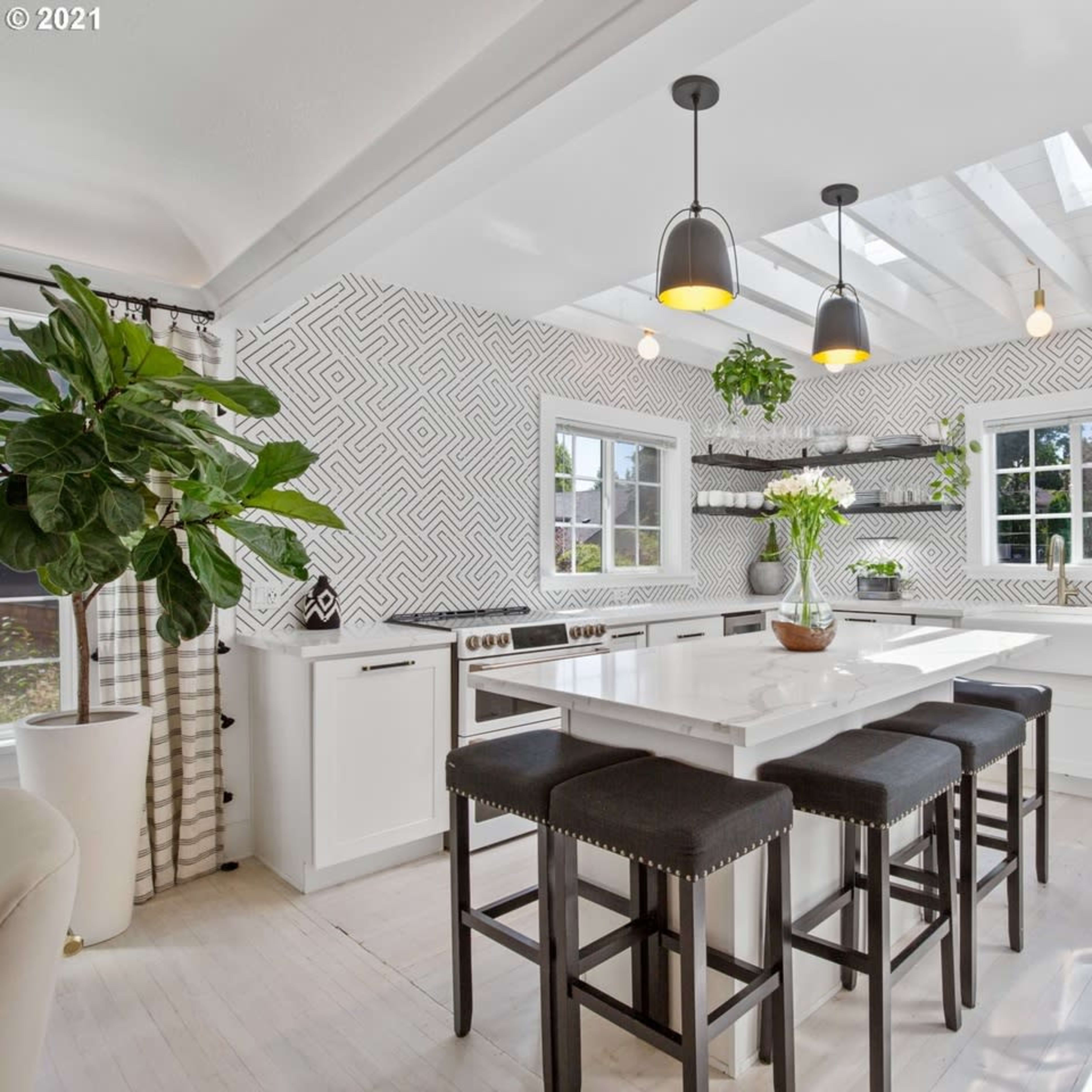 A bright kitchen features a white island with black stools, surrounded by plants and modern lighting, against a geometric-patterned backsplash.