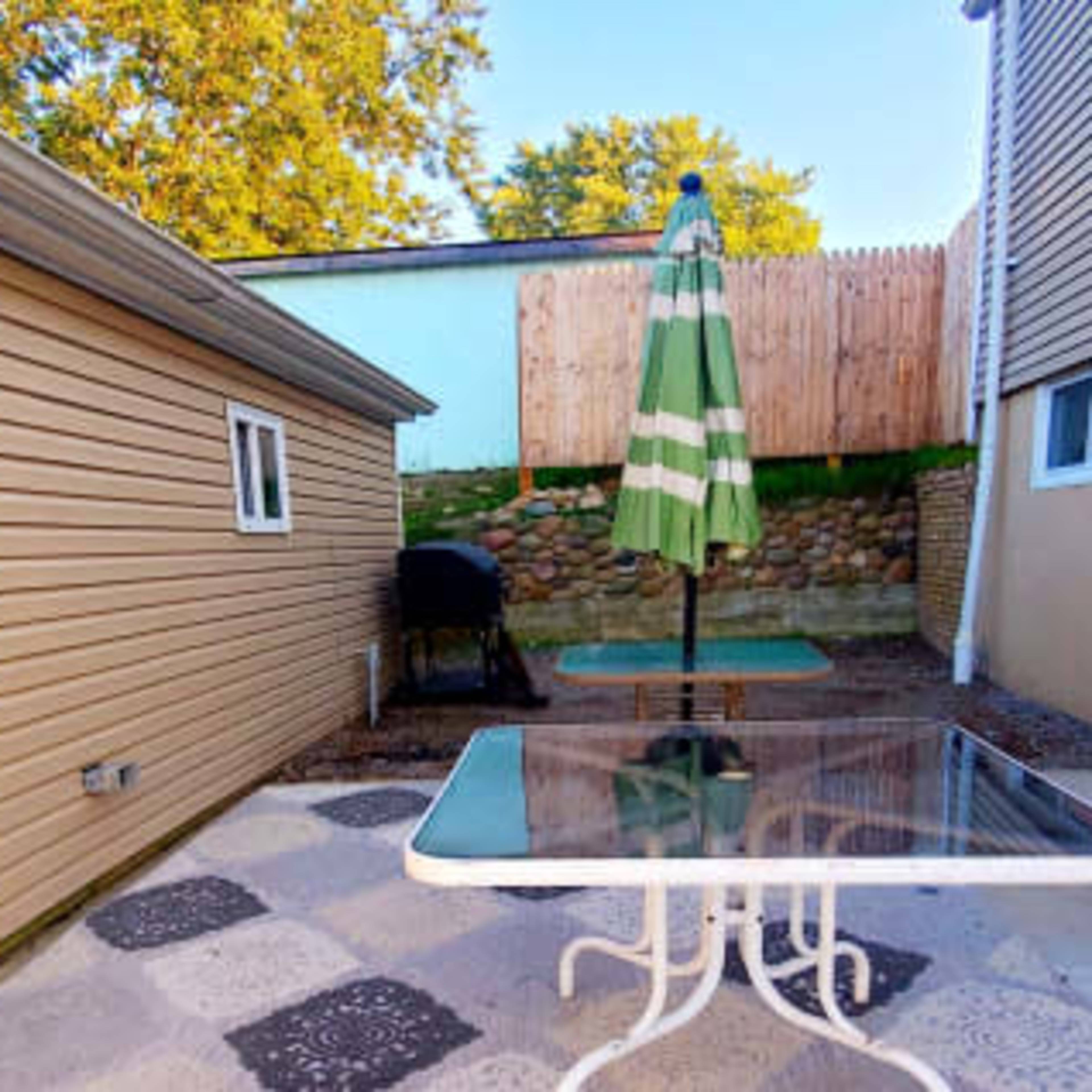 A patio area features a table with a glass top, a green and white striped umbrella, and a grill against a backdrop of two walls.