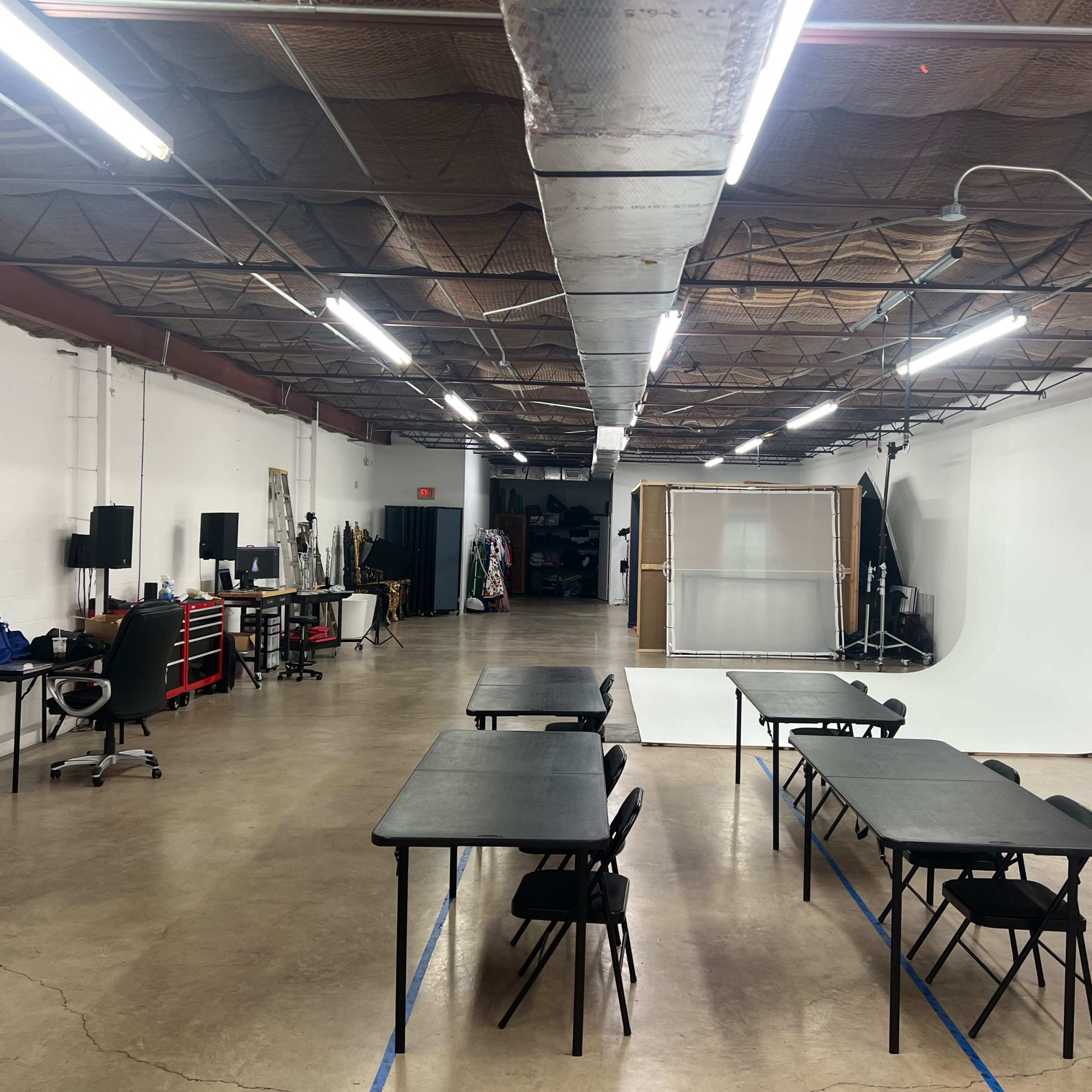 A spacious, well-lit studio with several tables and chairs arranged in the foreground and a white backdrop set up in the background.