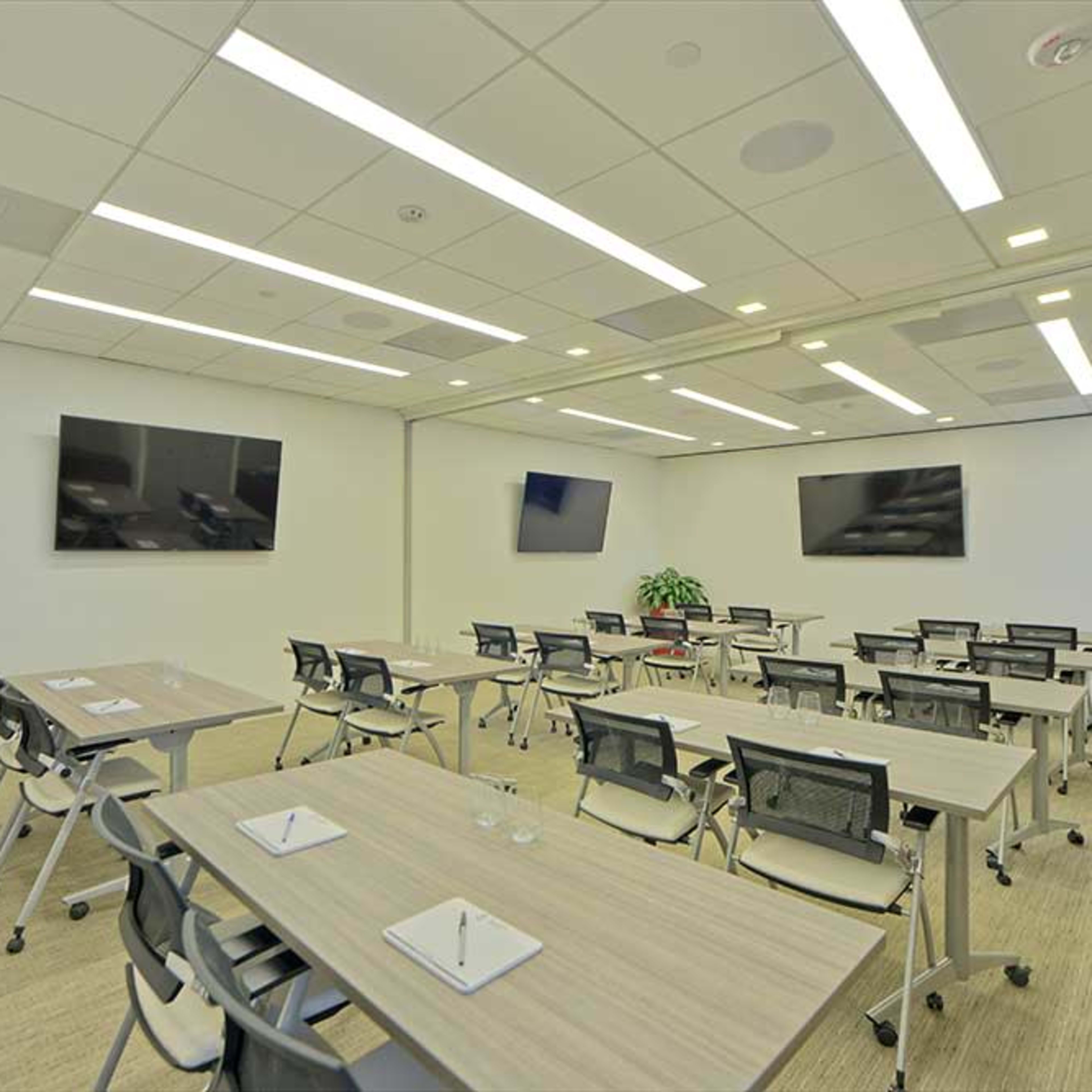 A modern conference room features multiple tables with chairs, two large screens mounted on the walls, and a potted plant in the corner.
