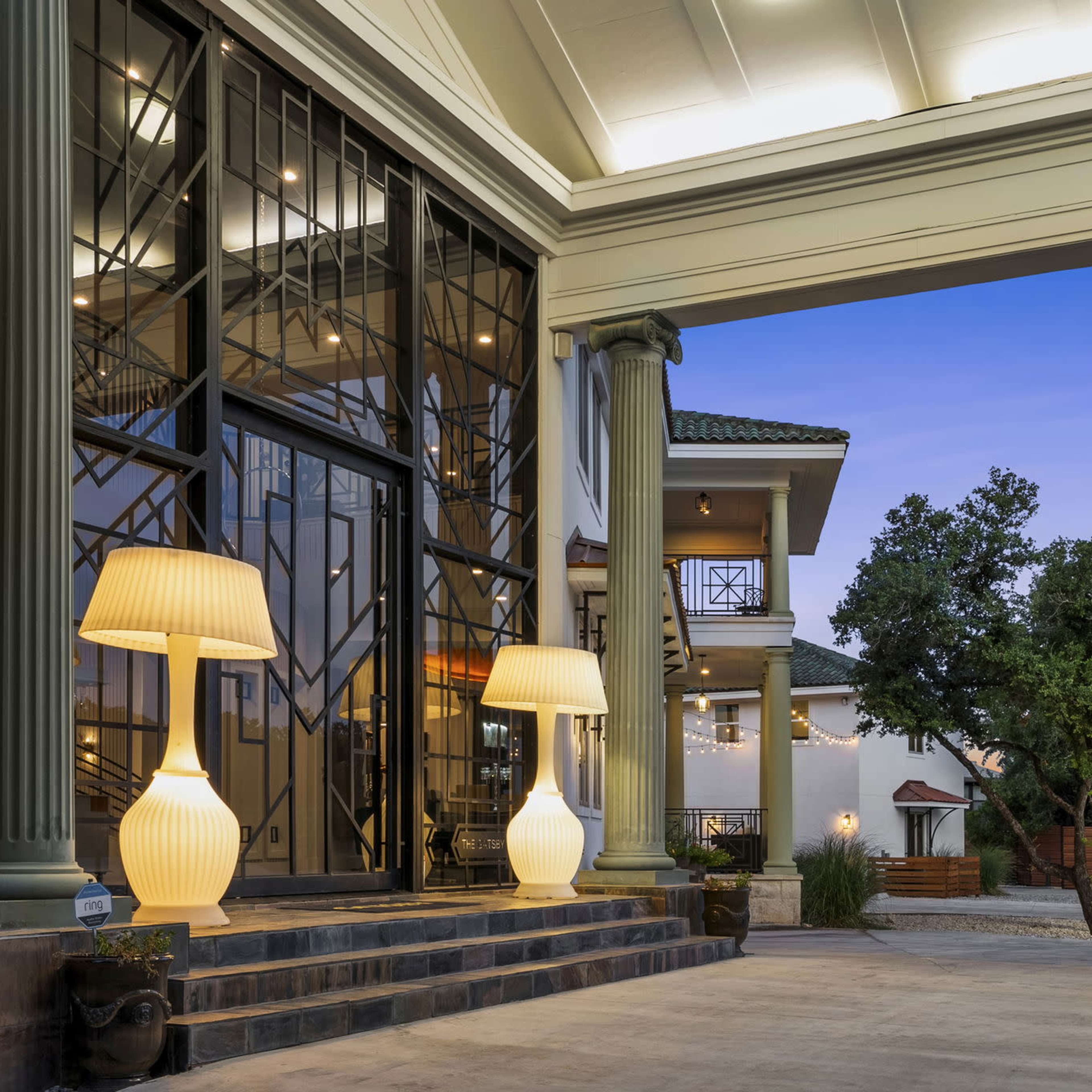 A large entrance with stylish columned architecture, featuring two illuminated lamps, and a view of a landscaped area with modern sculptures in the background.
