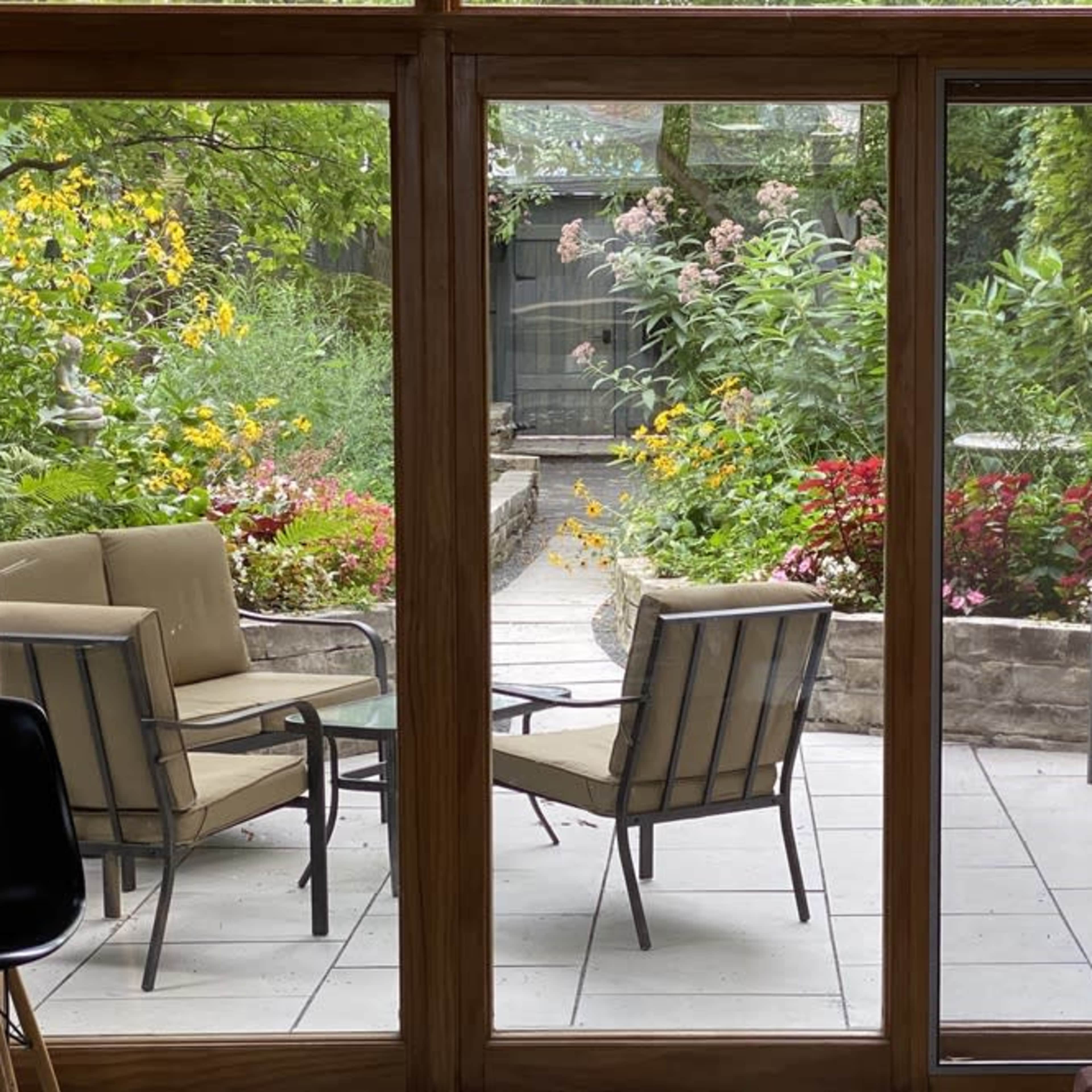 A patio area with four chairs and a small table is visible through large glass windows, surrounded by a garden filled with flowers and greenery.