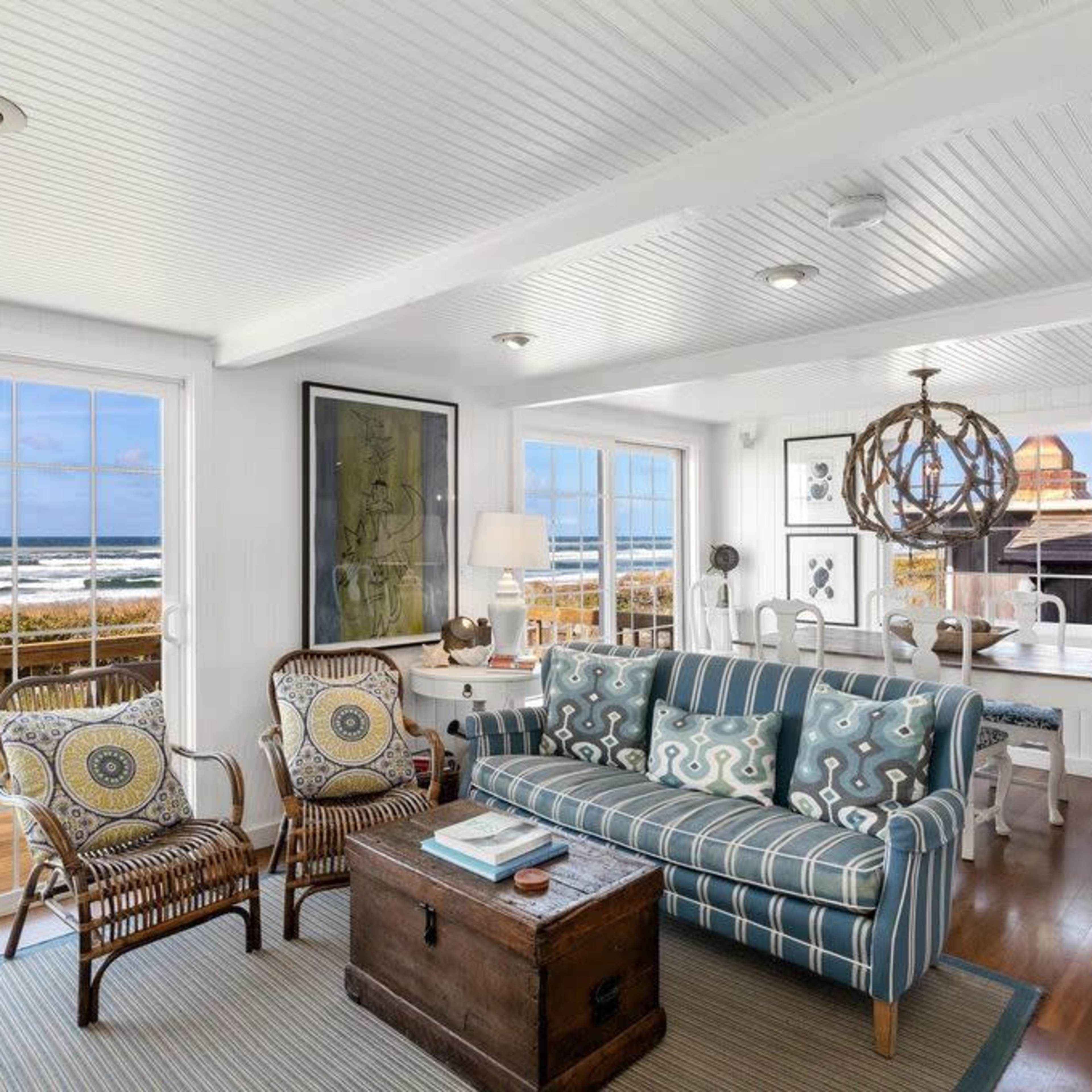 A bright, coastal living room with a striped sofa, wicker chairs, and large windows offering a view of the ocean.