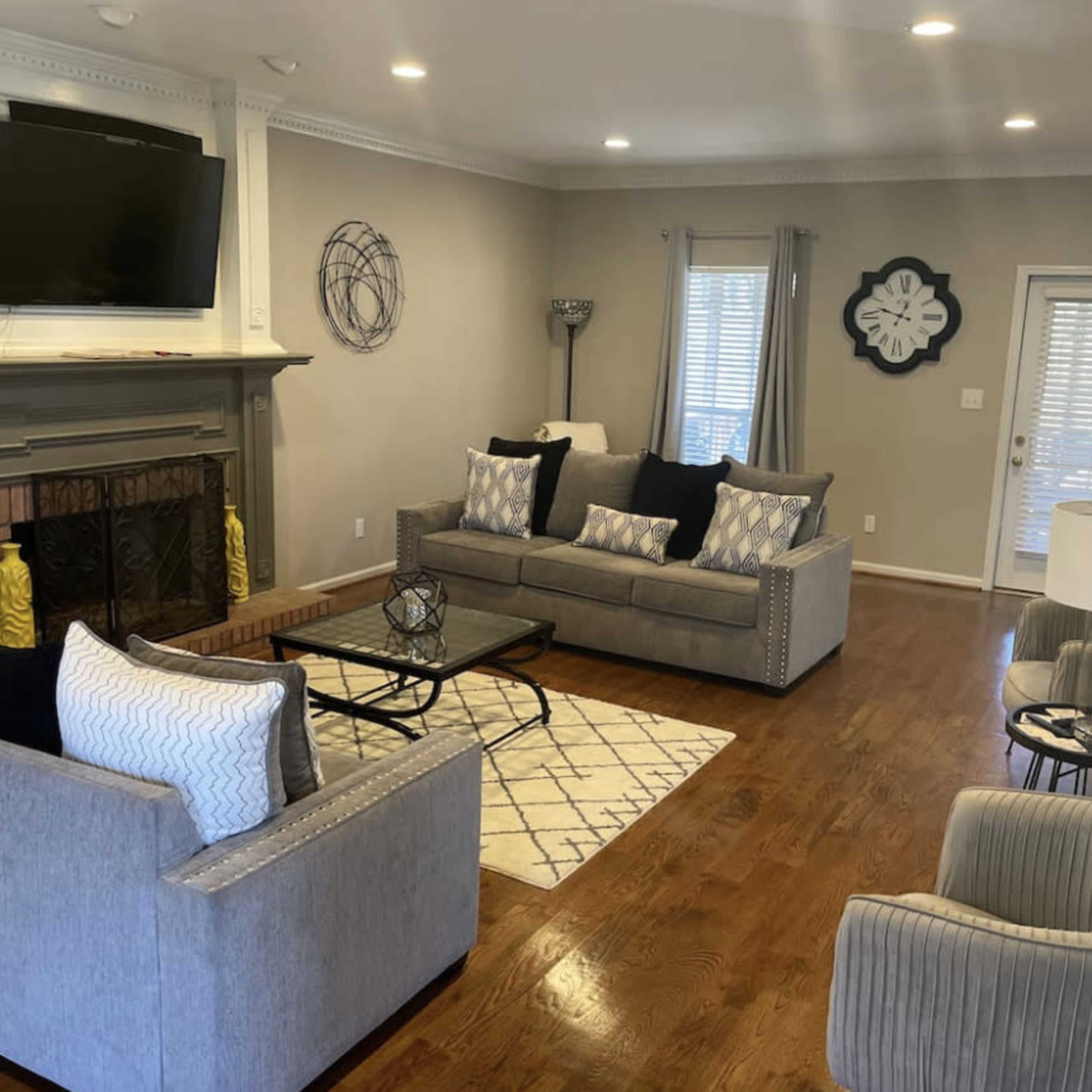 A living room features a gray sofa set, a coffee table on a patterned rug, a fireplace with a TV above it, and multiple windows providing natural light.