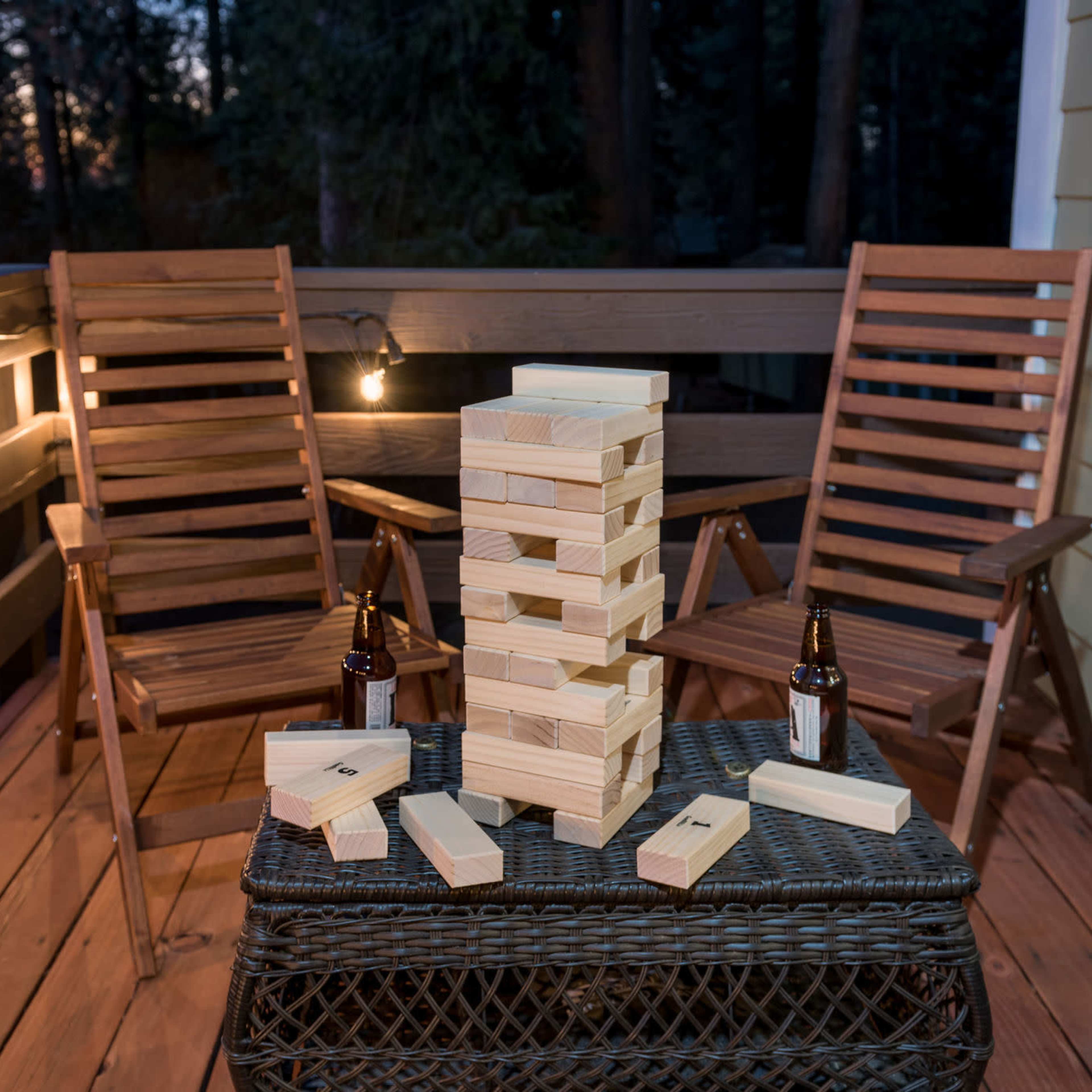 A set of wooden blocks is stacked in a tower on a table, surrounded by scattered blocks and two chairs on a dimly lit balcony.