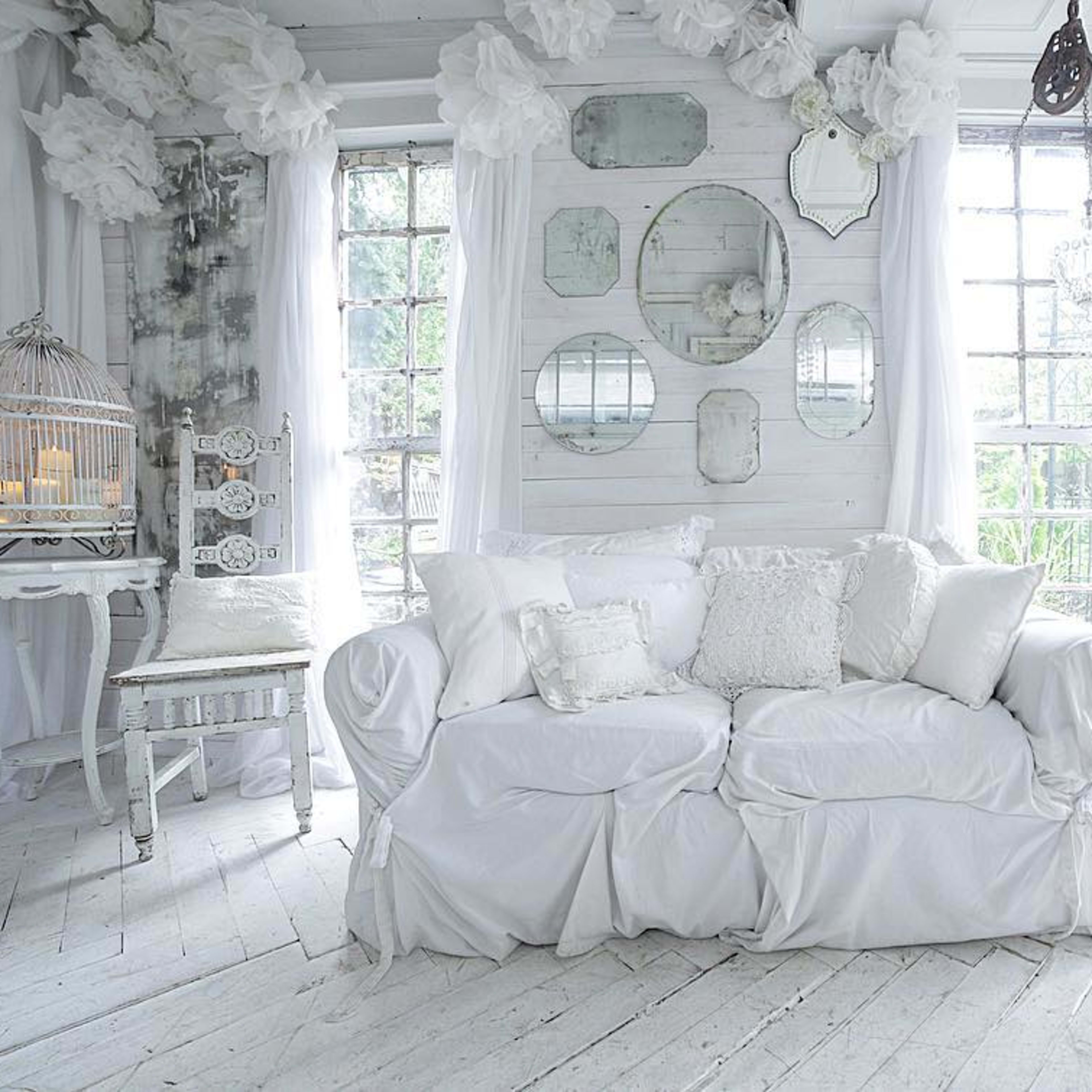 A bright, white living room features a large, draped couch, decorative mirrors on the wall, and vintage furnishings.