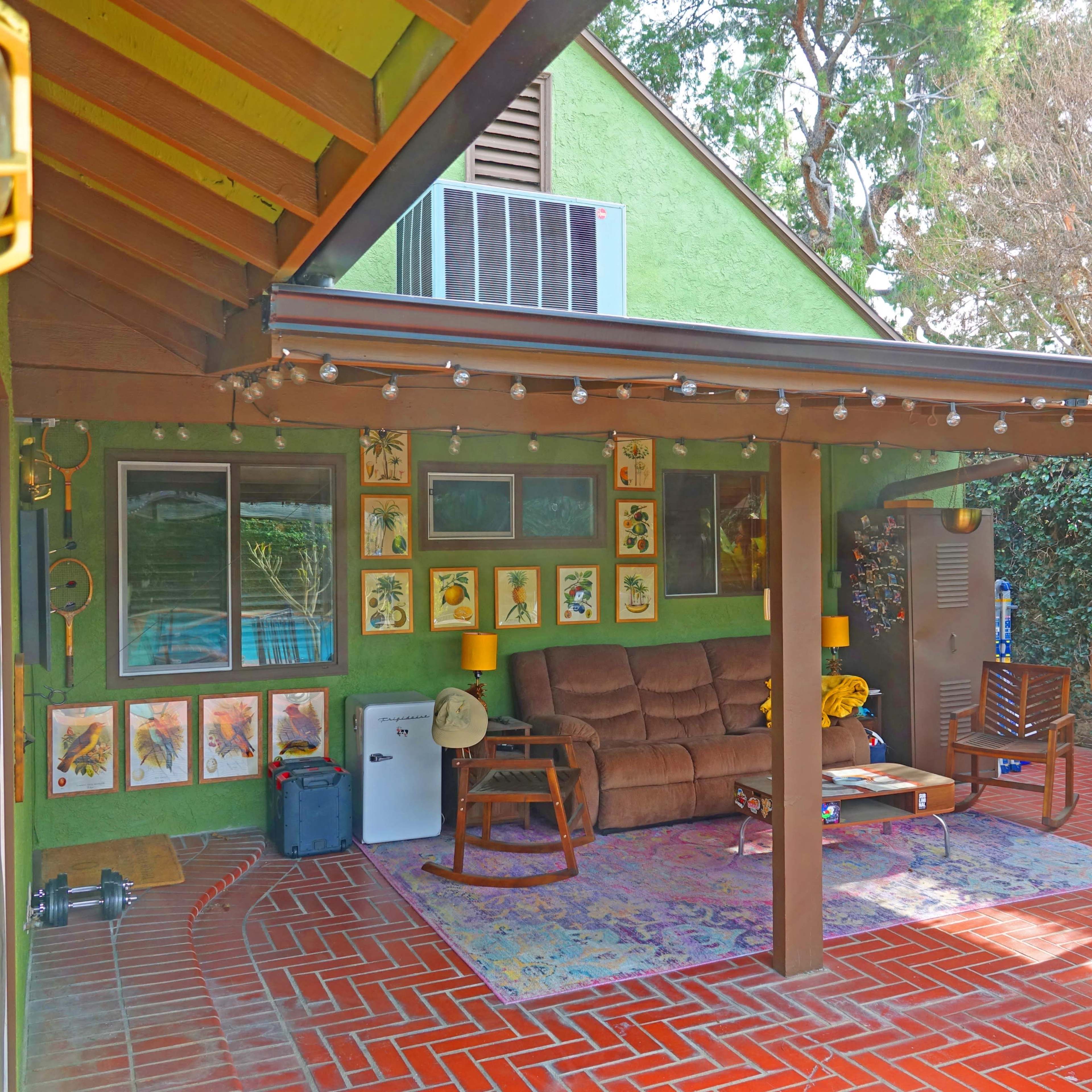 A covered patio area featuring a brown couch, various framed artworks on the walls, and a dining set on a patterned rug.