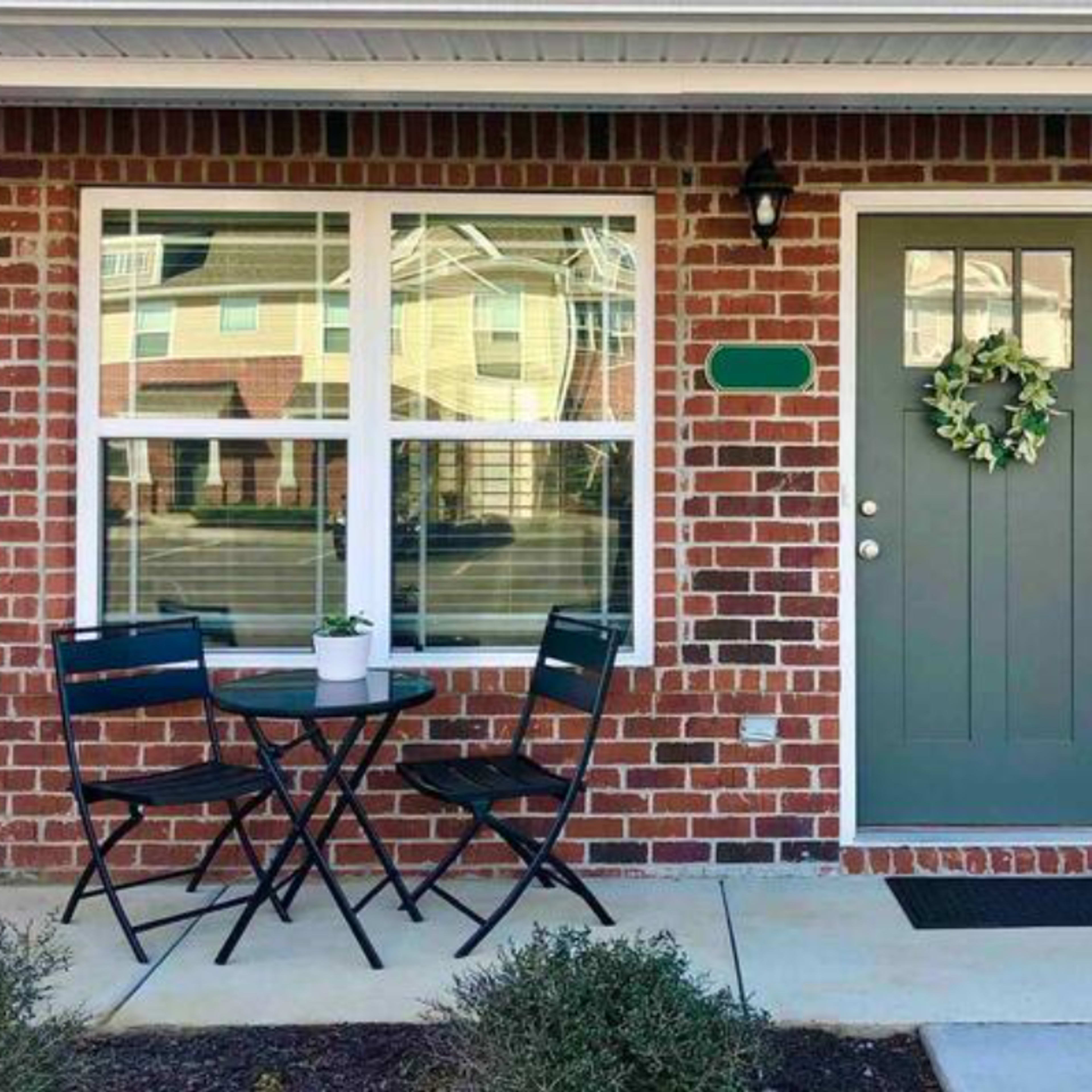A small outdoor seating area with a round black table and two chairs is set up in front of a brick house, next to a green door decorated with a wreath.