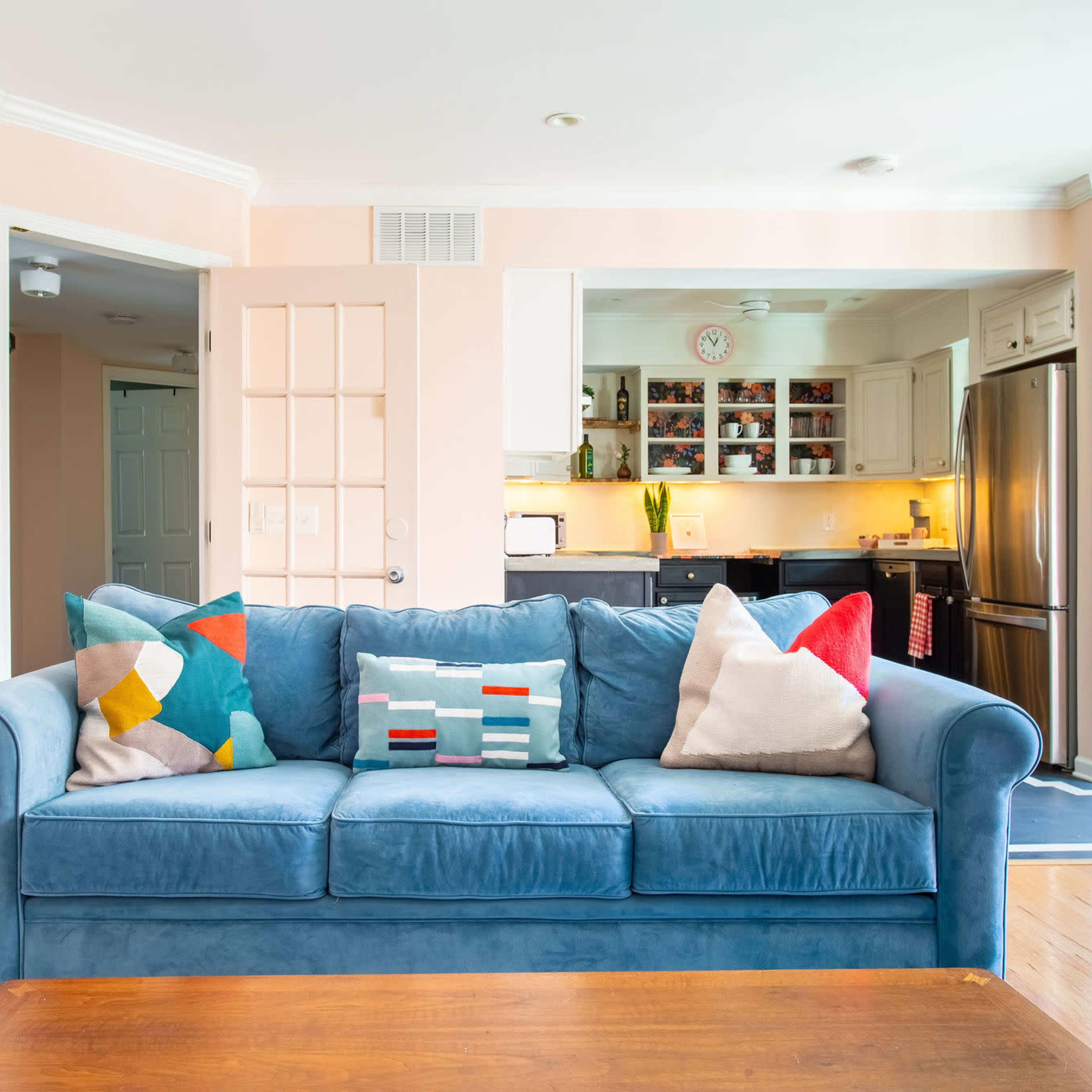 A blue sofa with patterned pillows is positioned in a bright living area that opens into a kitchen with wooden cabinets and appliances.